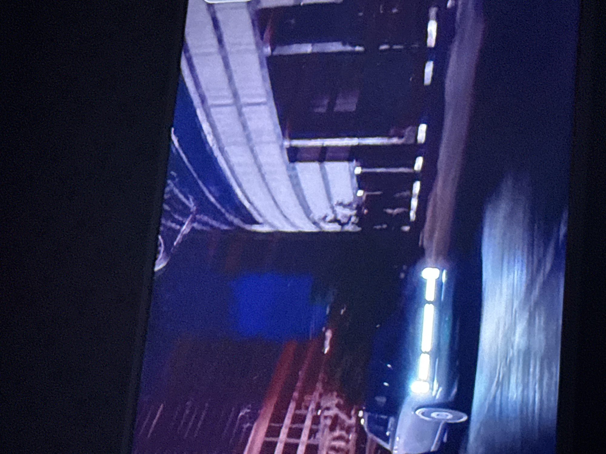 The image depicts a night scene in Mytishchi, Russia. An elevated metro line with a blue and white exterior is visible overhead. Below, a roadway or track is present, with a vehicle moving along it. The car's headlights are illuminated. The environment is dark, with limited ambient lighting.
