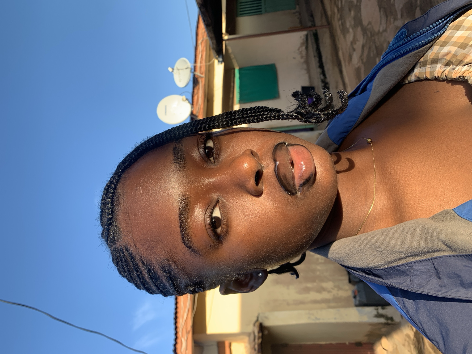 A close-up portrait of a young Black woman with dark skin, braided hair, and dark lipstick. She is looking directly at the camera, with a slightly open mouth. Her hair is styled in tight cornrows with a single braid falling over her left shoulder. She is wearing a blue and gray jacket over a checkered shirt, and a thin gold chain around her neck. The background appears to be an outdoor setting in a residential area, possibly in Rafin Par, Nigeria, with buildings and satellite dishes visible under a clear blue sky. The lighting suggests it is late afternoon, with the sun casting a warm glow on her face and skin.