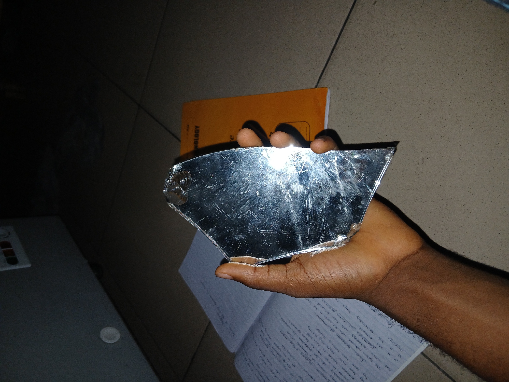 An indoor, close-up shot captures a person's hand holding a jagged piece of a broken mirror. The hand, with brown skin tone and some visible arm hair, firmly grasps the irregularly shaped, highly reflective object. The mirror shard is heavily scratched and reflects a bright light source, creating a strong glare that partially obscures the hand.
In the background, an orange book with the vertical text "NOLOGY" (likely part of "TECHNOLOGY") is visible. Below the hand, an open notebook with handwritten text is spread out on what appears to be a tiled floor. To the far left, a portion of a white or light-grey electronic device is partially visible. The overall lighting is somewhat dim, suggesting an indoor setting potentially in the evening or at night.
Visible text from the notebook includes phrases like "The system network was induced in the fo", "man", "xhnology", "extremely", "Cappeity.", and various numbers (e.g., "14", "155", "5150", "754"). An Arabic date "12 من في ١٨٩٧" (12th of in 1897) is also decipherable. The scene is likely set in a room in Lagos, Nigeria, as per the context.