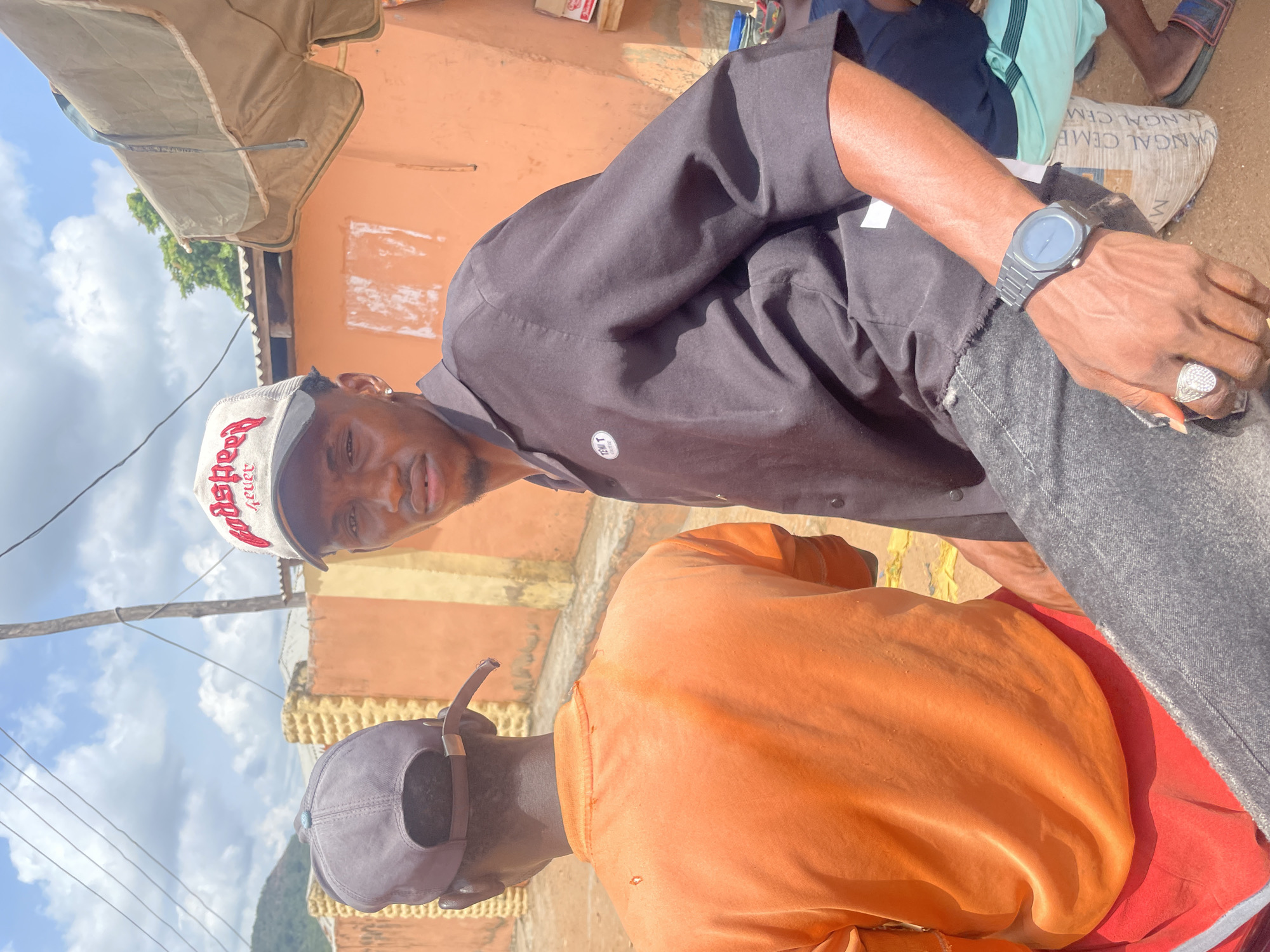 The image depicts two individuals outdoors in Lokoja, Nigeria. The first person, facing the camera, wears a white and red baseball cap, a black shirt, dark jeans, a watch on their left wrist, and a ring on their left hand. The second person, with their back to the camera, is wearing a gray baseball cap and an orange shirt. The background consists of an orange building, a blue sky with white clouds, overhead power lines, and a partially visible mountain. Another person is sitting on the ground with mint-colored pants.