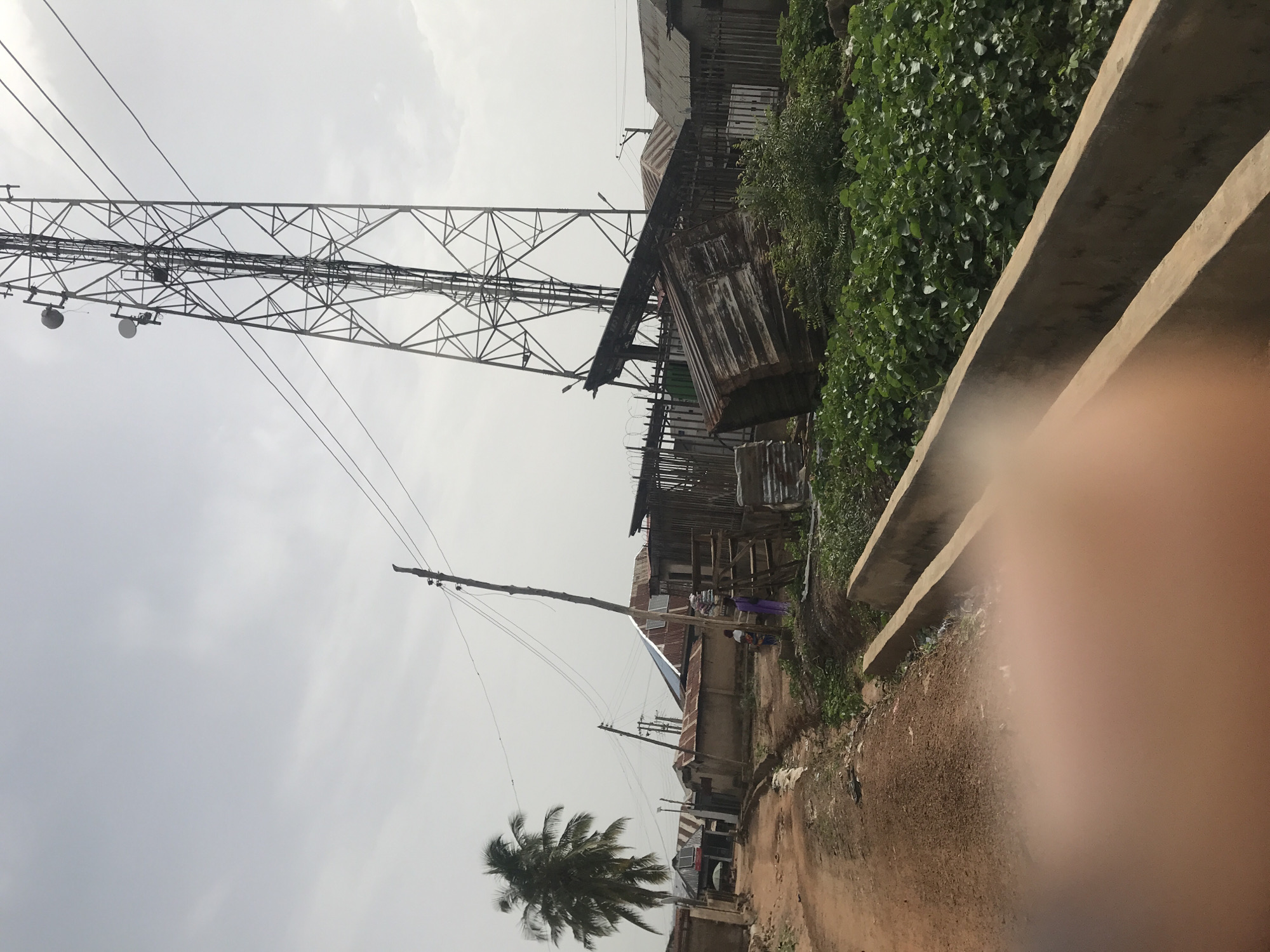 This outdoor image captures a semi-urban or rural street scene in Ilorin, Nigeria, under an overcast sky. The prominent feature is a tall, metal lattice communication or electricity tower on the left, adorned with several antennae and cables. To the right of the tower, a cluster of simple structures made from corrugated metal sheets and wood line a raised bank, some appearing worn or dilapidated.
A dirt road or path runs across the bottom and curves up the right side of the frame. Along this path, a low concrete barrier or curb is visible. Lush green foliage covers the bank alongside the structures. Towards the bottom center, a prominent palm tree stands, and several wooden utility poles with numerous overhead wires crisscross the sky, indicating local infrastructure.
Two individuals are partially visible in the mid-ground near the structures. One person is wearing purple attire, and the other is in a white top and patterned skirt/wrapper, seemingly going about their daily routines. The general atmosphere suggests an ordinary daytime scene, possibly late morning or afternoon, given the diffuse light from the cloudy sky. No specific event is taking place, and no text is discernible. A blurry, flesh-toned object (likely a finger) obscures the bottom right corner of the image.