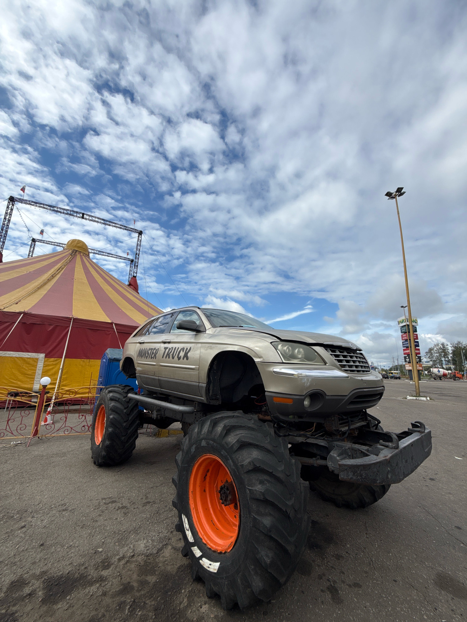 A tan colored monster truck is parked in an open lot on a cloudy day. The truck is modified with oversized tires, orange rims, and a raised suspension. The word "MONSTER TRUCK" is visible on the side of the truck. In the background, a large red and yellow striped circus tent is set up. Various flags are visible on poles and on the tent. A tall light pole stands in the background to the right. The ground is paved with asphalt. This scene is located in Rusanovo, Russia.