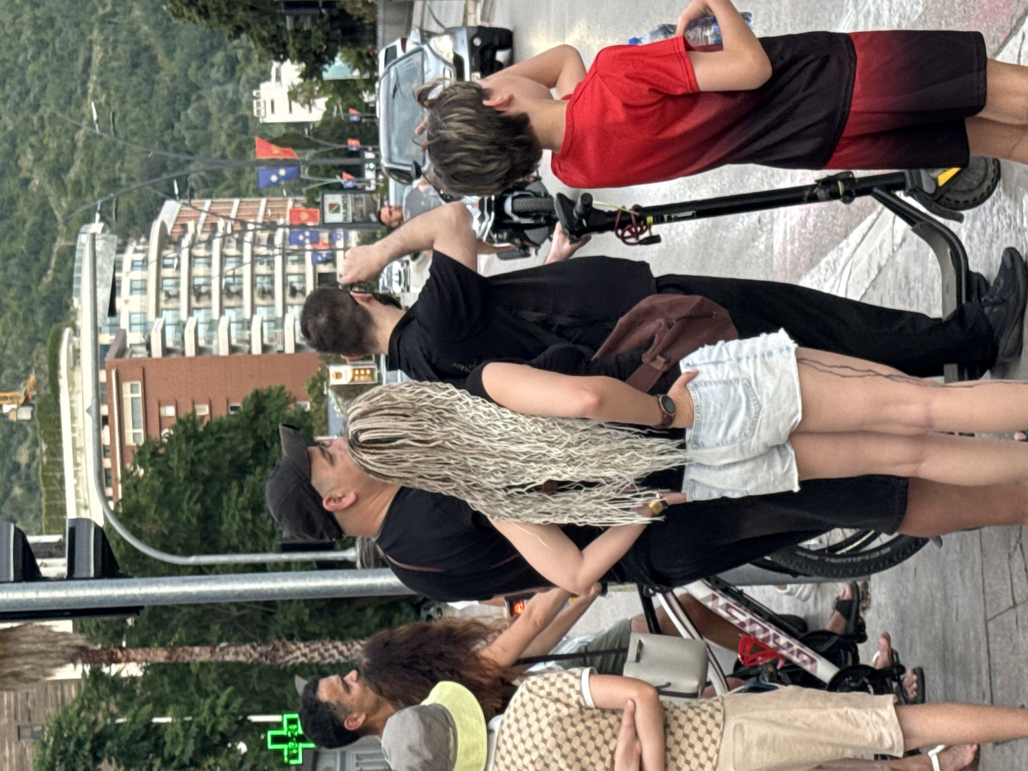 In Budva, Montenegro, a group of people are gathered on a sidewalk near a busy street. A traffic light stands tall, a palm tree is visible to its left, and a building with many floors and balconies dominates the background, with a tree-covered hill behind it. On the sidewalk, a man in a black baseball cap stands next to a woman with long blonde dreadlocks. To their left, a man in a black t-shirt is looking up. In the foreground, a young boy in a red shirt stands with a scooter. A white bicycle is parked nearby. The street is wet, suggesting recent rain, and cars are visible in the background. The overall atmosphere is that of a pedestrian area within a city.