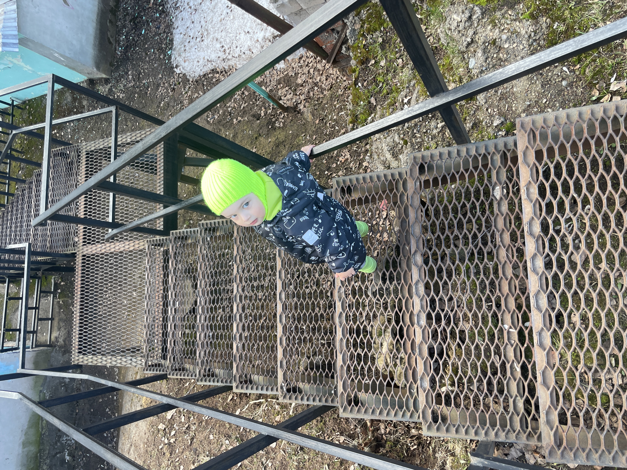 A small child in a black and white printed snowsuit and bright green hat and boots stands on a metal grate staircase. The staircase is part of an industrial structure in an outdoor area. Vegetation and loose debris are visible through the grates. Some snow remains on the ground nearby. The photo appears to be taken from above, looking down at the child. The location is Izhevsk, Russia.
