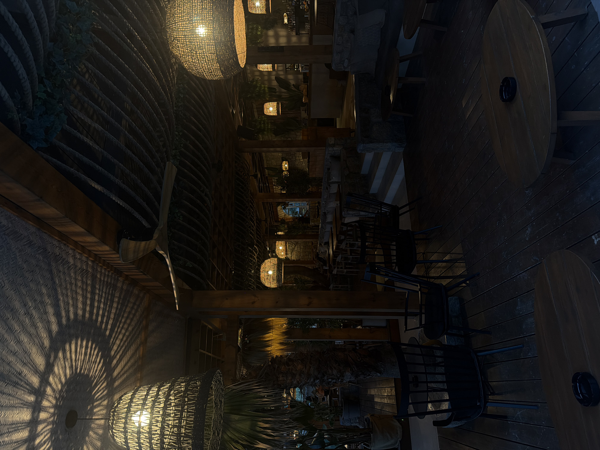 This is an outdoor scene at night, likely a restaurant or bar, with warm ambient lighting. Large, woven pendant lamps hang from the wooden-beamed ceiling, casting intricate shadow patterns. The ceiling itself is decorated with rope and some greenery. In the foreground, there are dark wooden tables and chairs, suggesting seating for patrons. The floor is also made of dark wood planks. Beyond the immediate seating area, there's a glimpse of a bar or counter area with more seating and stone-built structures. The overall atmosphere appears relaxed and inviting. While the prompt mentions Sithonia Municipal Unit, Greece, this specific visual information cannot be determined from the image alone.