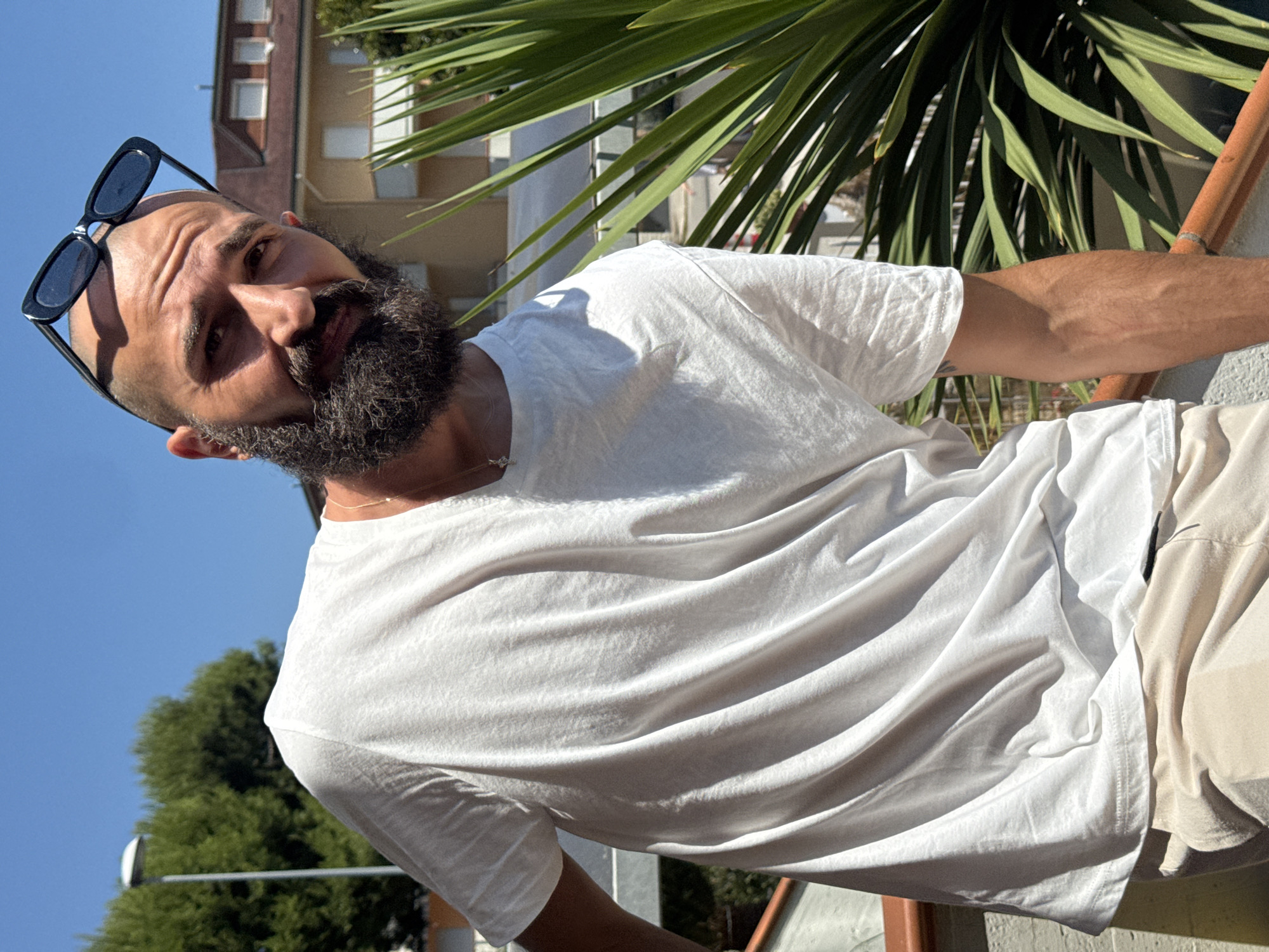 A man with a beard and sunglasses is shown from the chest up. He is wearing a white t-shirt and tan shorts. He is outdoors, with a palm tree in the foreground and a building in the background. The sky is clear and blue, suggesting it is daytime and sunny. The man is looking at the camera with a slight smile. The photo appears to be a selfie, taken by the man himself. The scene suggests a casual, outdoor setting, possibly a vacation or a relaxed day. The sun is casting shadows, indicating it is likely midday or afternoon. There is no visible text in the image.