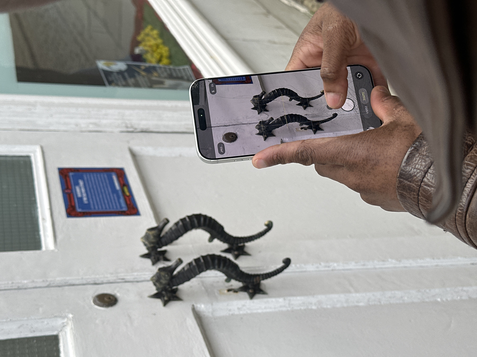 A person is holding an iPhone and taking a photo of two metal, seahorse-shaped door knockers on a white door. The door also has a glass panel with a blue plaque above it. The setting is likely a building in the Folkestone and Hythe District, United Kingdom, given the style of the door and door knockers. The person is wearing a brown leather jacket.