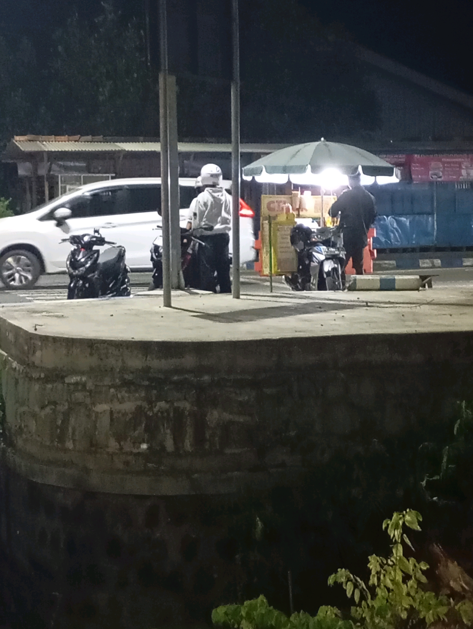 The image depicts a nighttime scene at a street food stall in Pacitan, Indonesia. A white car is parked to the left, partially obscured by a concrete barrier in the foreground. Two individuals, one wearing a white helmet and a light-colored hooded jacket, and another in dark clothing, stand near motorcycles. A food cart with an umbrella is visible in the background, illuminated by bright lights. The surrounding environment appears to be a paved area with some vegetation visible on the right side of the image. The overall atmosphere is one of a typical street scene at night, possibly near a road or parking area.