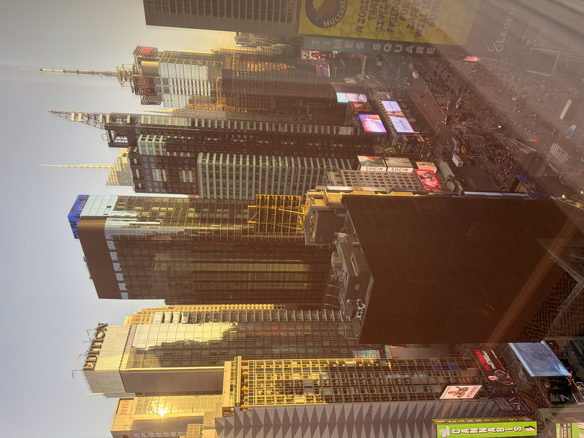 The image depicts an elevated view of Times Square in New York City. Numerous skyscrapers and buildings surround the square. Digital billboards display advertisements. A dense crowd of people are milling about on the streets below. The sky is a pale blue, indicating either early morning or late afternoon light. The "EDITION" hotel and a billboard for "CANNABIS" are visible. A sign displays "RIU," likely another hotel or brand. A "SHEIN" advertisement is also visible.