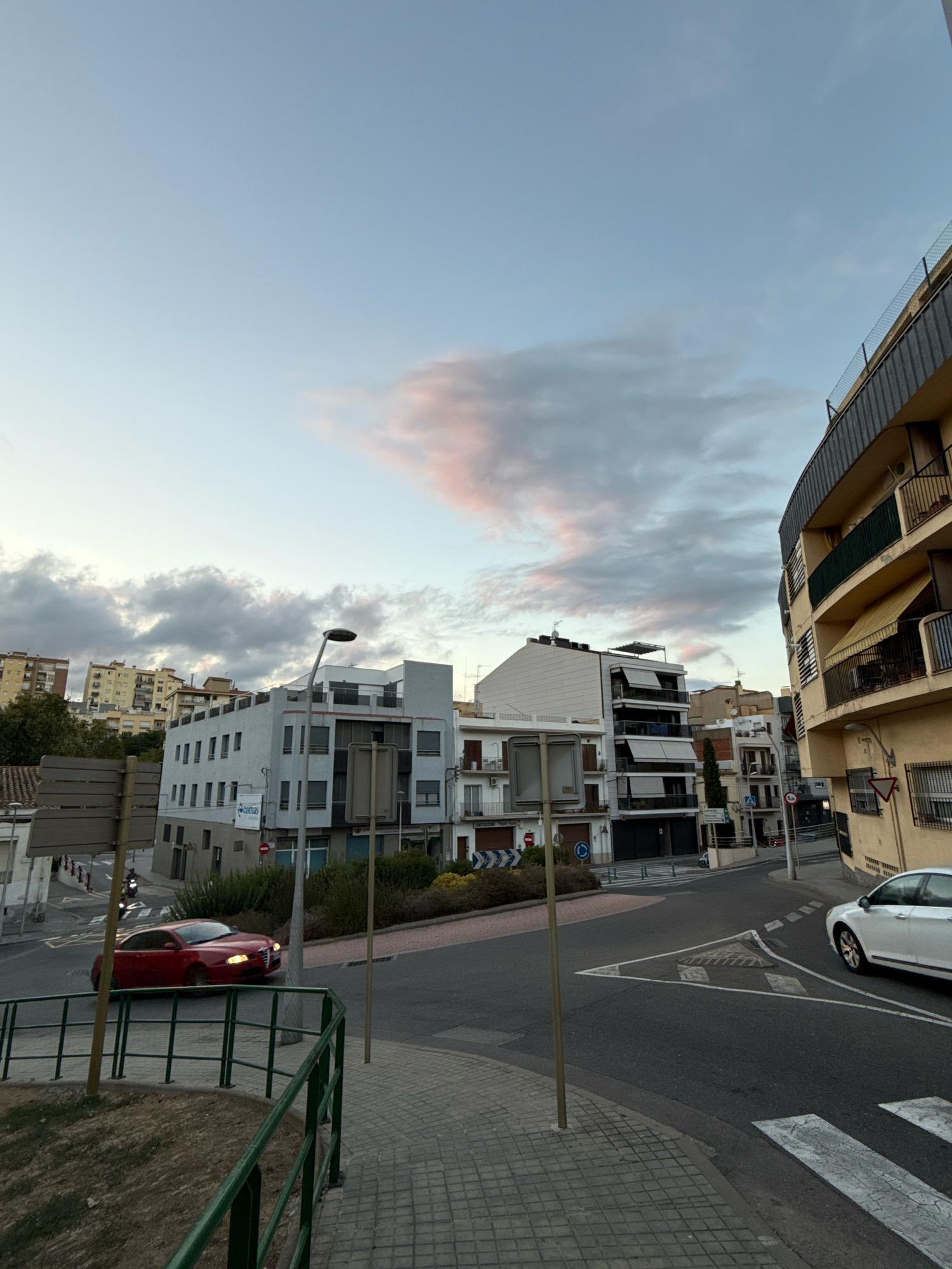 The image depicts a street scene in Blanes, Spain, at what appears to be dusk. In the foreground, a paved sidewalk with a green metal railing lines the edge of the street. A red car is visible driving on the asphalt road, heading towards the left of the frame. Further down the street, several multi-story buildings with balconies are present. The sky above is partly cloudy with hues of pink and orange, suggesting the setting sun. A street lamp stands on the right side of the road, illuminating the scene. In the background, more buildings are clustered on a hillside.