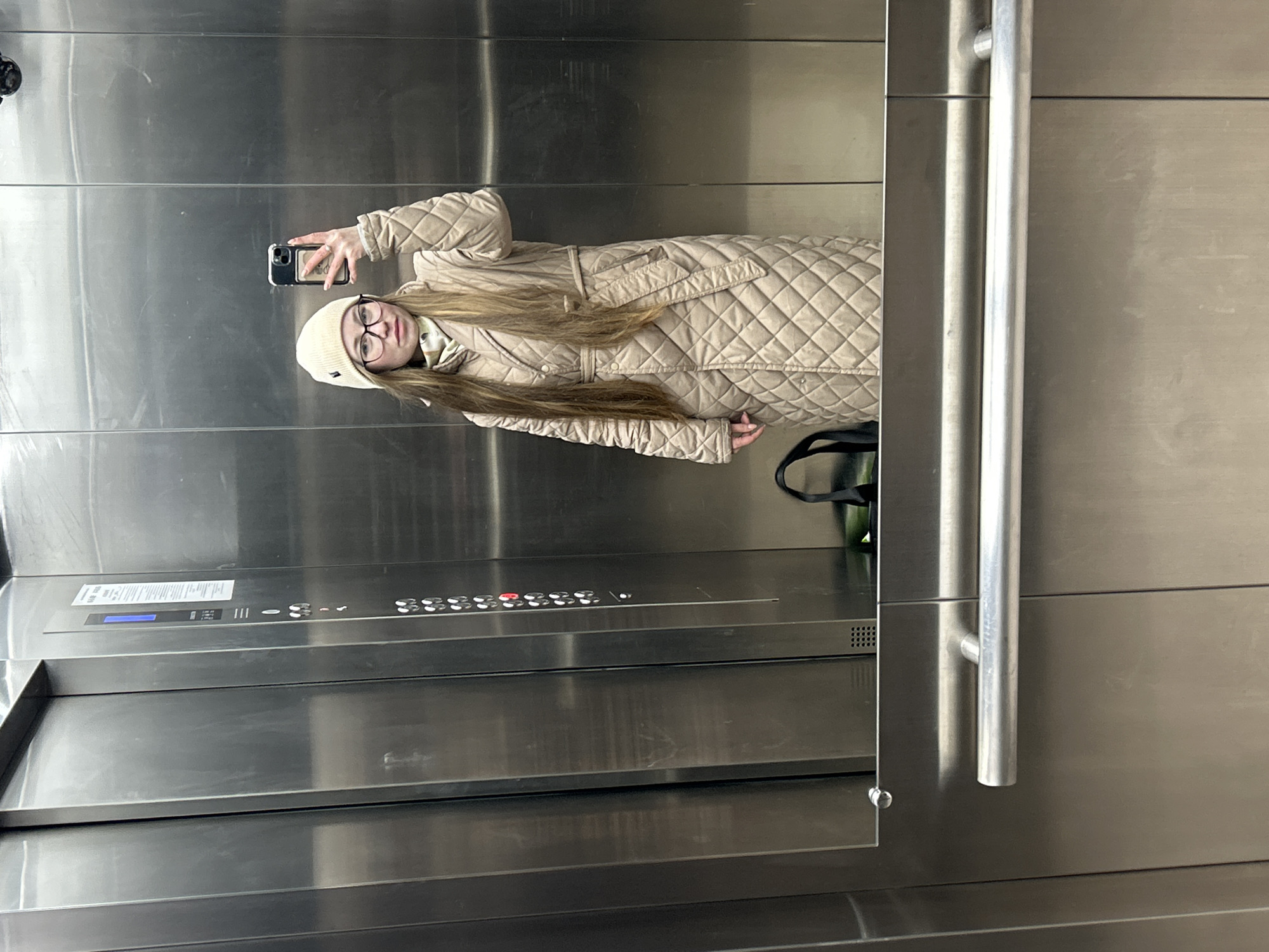 A young woman is captured in a mirror selfie inside what appears to be an elevator in Saint Petersburg, Russia. The image is oriented sideways, with the woman standing upright relative to the frame. She holds a dark-colored smartphone with a light-colored case in her right hand, taking the picture of her reflection.
The woman has long, straight brown hair that cascades down her front. She is wearing a light beige or cream-colored quilted puffer coat, which appears to be long and belted, and a matching knitted beanie. She also wears dark-rimmed glasses and has a neutral expression on her face. A subtle ring is visible on her right hand.
The elevator interior is predominantly made of brushed stainless steel. To the left of the woman, the elevator control panel is visible, featuring a digital display that shows blue numbers, likely indicating the current floor. Below this display are various buttons, some of which are illuminated in red. Further to the right, a horizontal silver handrail is mounted on the wall. A dark bag with green accents can be seen partially reflected near the woman's left side. The overall lighting suggests an indoor, artificial light source. No specific time of day or weather conditions can be discerned from the image.