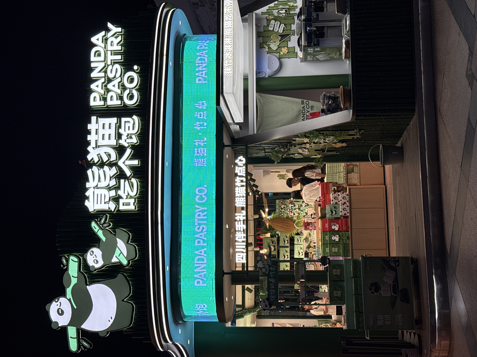 The image shows the exterior of "Panda Pastry Co." in Jinjiang District, China. The store has a vertical sign with the company name in English and Chinese, along with panda illustrations. A digital screen displays the same information with added details like "熊猫礼·竹点心" (Panda Gift, Bamboo Desserts) and "四川伴手礼 熊猫竹点心" (Sichuan Souvenir, Panda Bamboo Desserts). The store's interior is visible, featuring bamboo-themed decorations, shelves with products, and people inside. A sign reads "熊猫米团 ≥23%" (Panda Rice Ball ≥23%) and promotional material mentioning "抹竹冰淇淋 熊猫吃不停" (Bamboo Ice Cream, Panda Can't Stop Eating). Additional text includes "饱饱的春竹季" (Full Spring Bamboo Season) and "9.9" with a panda image.