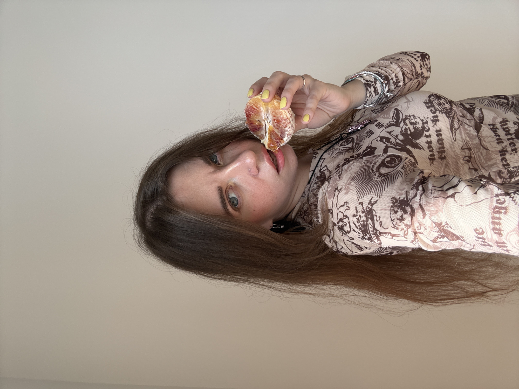 A young woman with long brown hair is holding a halved blood orange near her lips. She is wearing a patterned beige and brown long-sleeved shirt. Her nails are painted yellow. She is wearing a ring on her left hand and bracelets on her right wrist. The background is a plain beige wall. The photograph appears to have been taken from a slightly low angle. There is no indication of the location being Moscow, Russia.