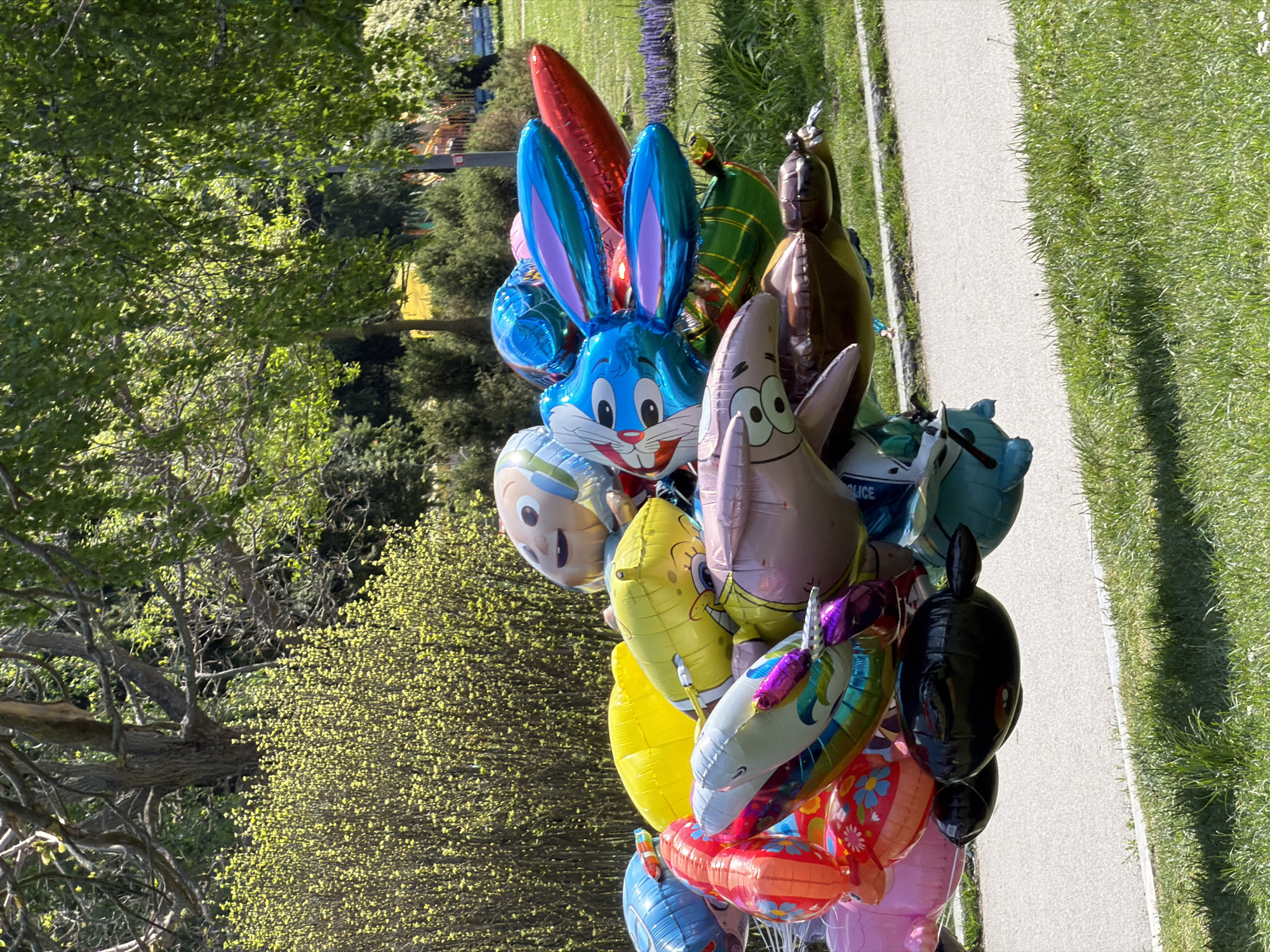 A collection of character balloons is positioned near a sidewalk and grass. The balloons include Bugs Bunny, SpongeBob, Patrick Star, a unicorn, and what appears to be a horse, a police car and other unidentifiable character balloons. The scene includes greenery and trees in the background. The location is potentially in Sofia, Bulgaria.