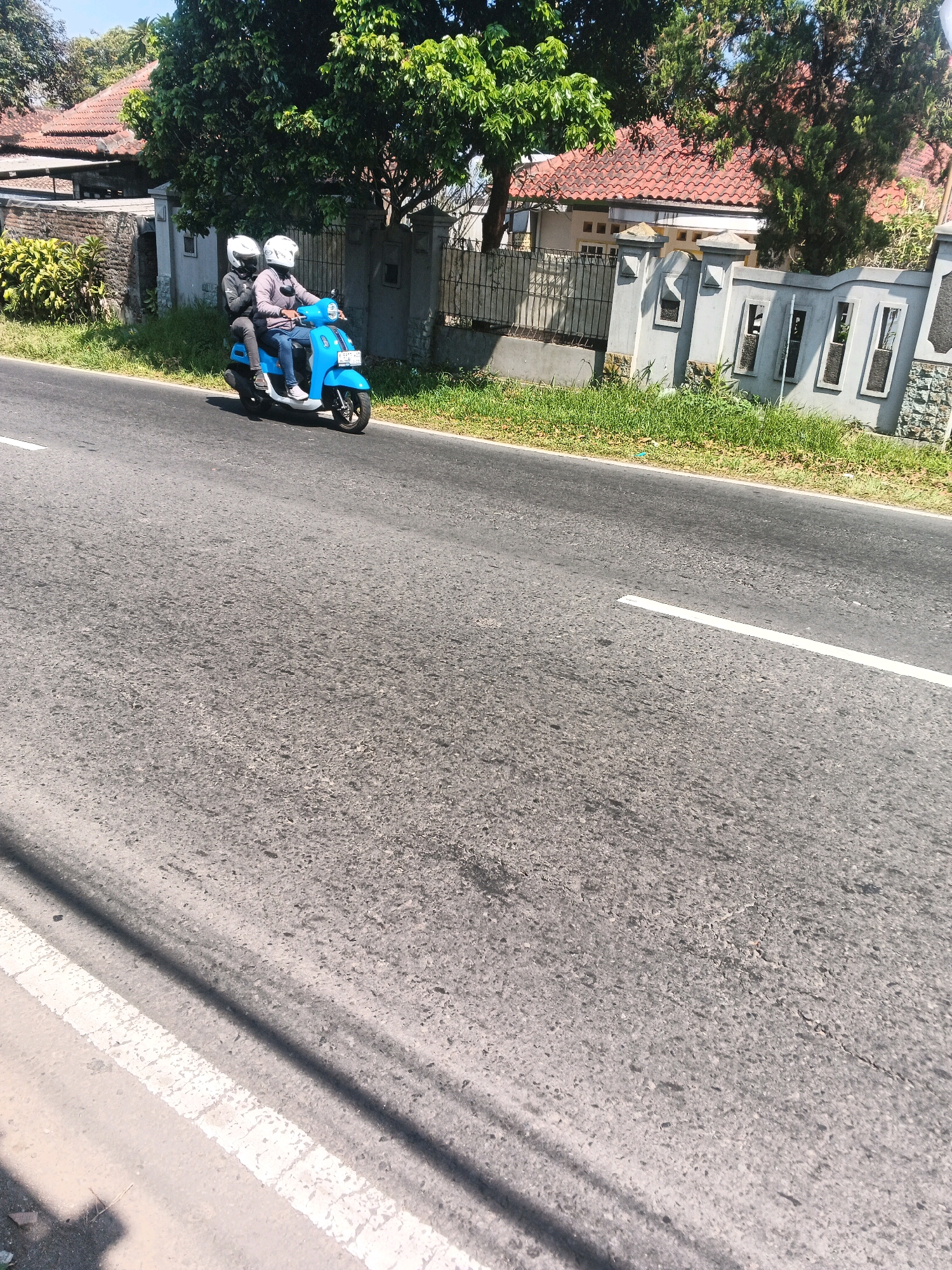Two individuals wearing helmets are riding a bright blue scooter on a paved road. The person in the front is driving, while the person in the back is a passenger. They are positioned in the middle lane of a two-lane road, which appears to be a street in Bandungan, Indonesia, given the presence of houses and tropical vegetation in the background. The houses have terracotta tile roofs and are situated behind a grey concrete wall with decorative rectangular openings. Trees with dense green foliage are visible on either side of the road. The road surface is asphalt, with a white line marking the lane. The lighting suggests it is daytime, with clear skies overhead.