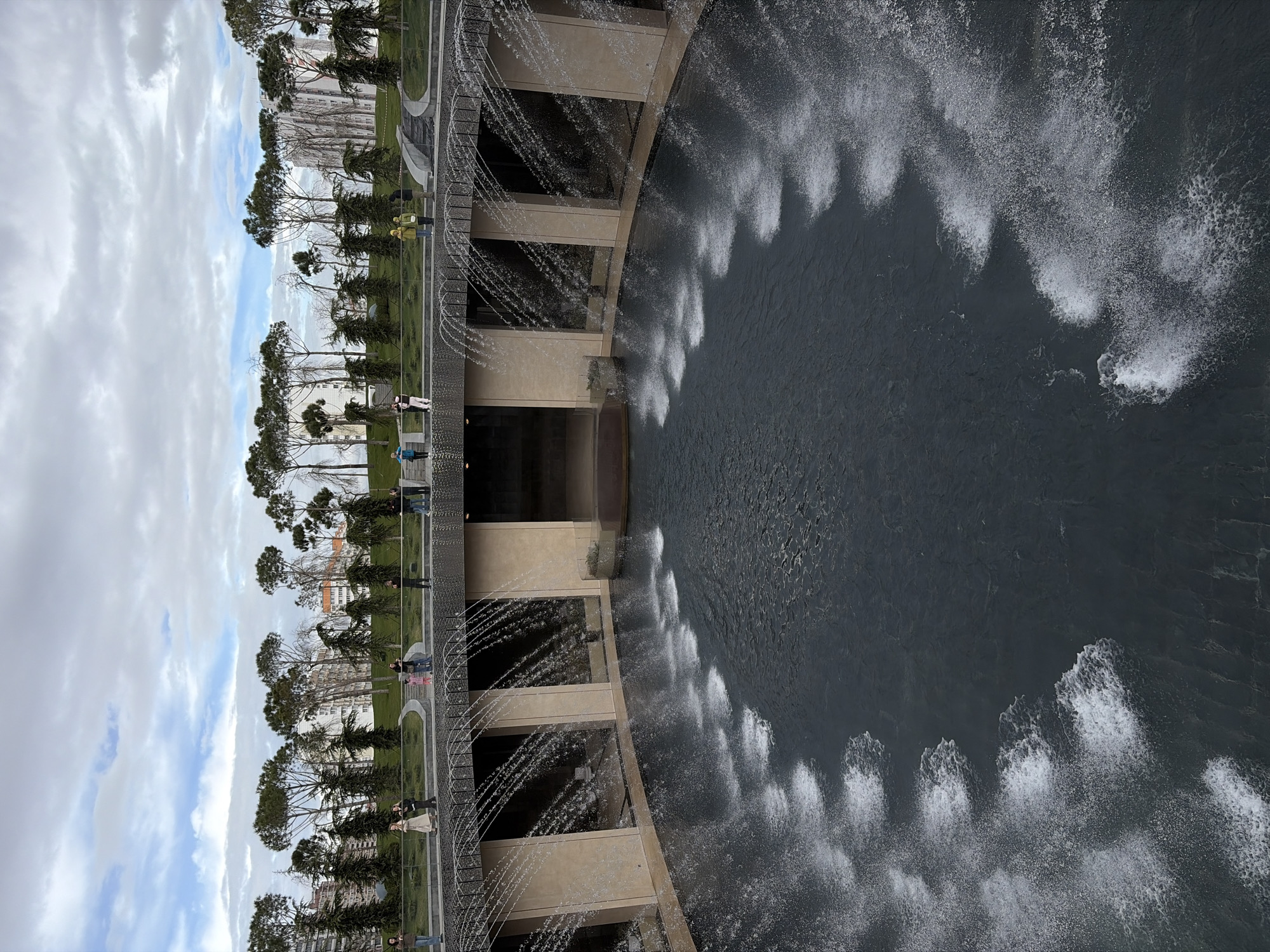 This photograph shows an artificial pond or reservoir in the city of Krasnodar, Russia. The viewpoint is from above, oriented at a 90-degree angle. Water cascades from a series of tiered, rectilinear concrete structures into the pond below. The water creates a series of prominent white sprays against the dark water. The reservoir is bordered by a pedestrian walkway with a dark metal railing. On the other side of the walkway, a narrow strip of grass is planted with small trees, suggesting a park-like setting. Figures of people can be seen walking along the walkway. The sky is partly cloudy with some blue patches visible.