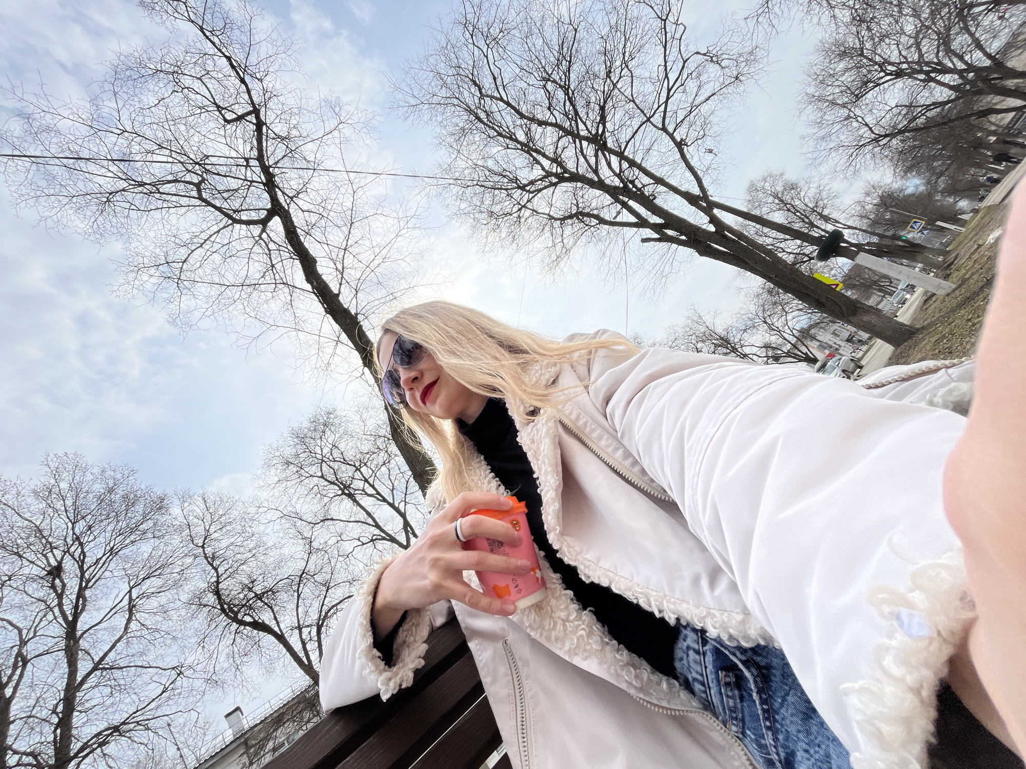 A low-angle, eye-level shot captures a woman sitting outdoors in what appears to be a park or public area in Chaikovsky, Russia. The trees are bare, suggesting it is either late autumn or early spring. The sky is overcast with some patches of blue visible.
The woman is fair-skinned with blonde hair and is wearing sunglasses, a black turtleneck sweater, blue jeans, and a white jacket with a fur collar. She is holding an orange paper cup.
Behind her and around her are many bare trees. In the distance, some buildings and street signs are visible. She is sitting on a wooden bench. The angle of the shot includes part of her arm and hand extending towards the camera on the right side of the frame.