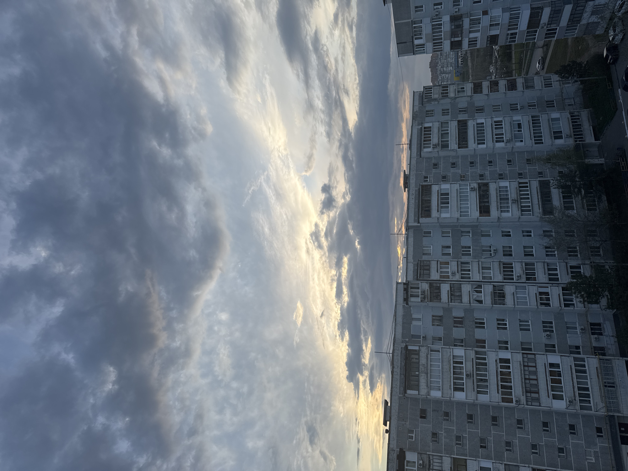 The image shows a multi-story apartment building in Kazan, Russia, under a cloudy sky at dusk or dawn. The building's facade is grey, with many windows and balconies. The sky is filled with dark grey and white clouds, and the sun is partially visible behind the clouds, creating a golden glow. A few trees and cars can be seen in the background around the building.