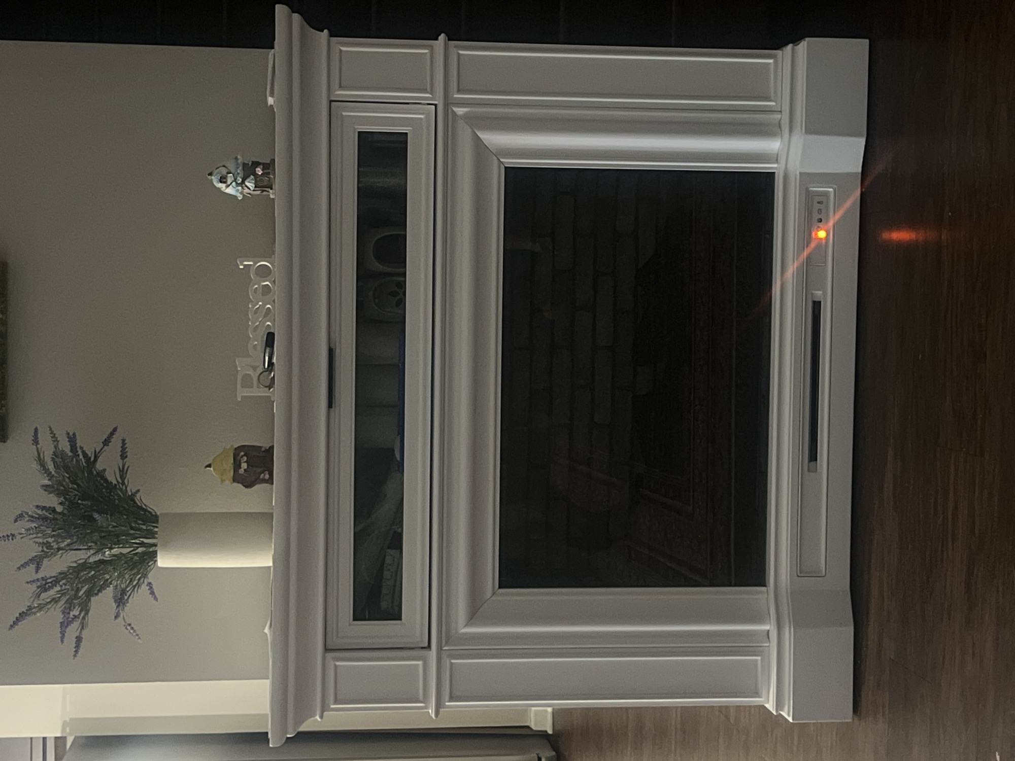 This is a head-on, medium shot of a white electric fireplace with a glass-fronted firebox, set against a neutral wall. The fireplace has a decorative mantelpiece on top, adorned with a white vase filled with artificial lavender, two small decorative figures resembling gnomes or whimsical houses, and a silver word art piece spelling "Blessed" with a key lying next to it. The firebox below the mantelpiece has a glass front revealing a brick interior and a dark floor. Below the glass, there is a control panel with a red indicator light illuminated, suggesting the fireplace is on. The fireplace sits on a dark wood-plank floor. The background is plain and light-colored, with a slight shadow cast on the left side, suggesting a light source from the left. There are no people visible in the image. The location of this image is not determinable from the visual information provided.