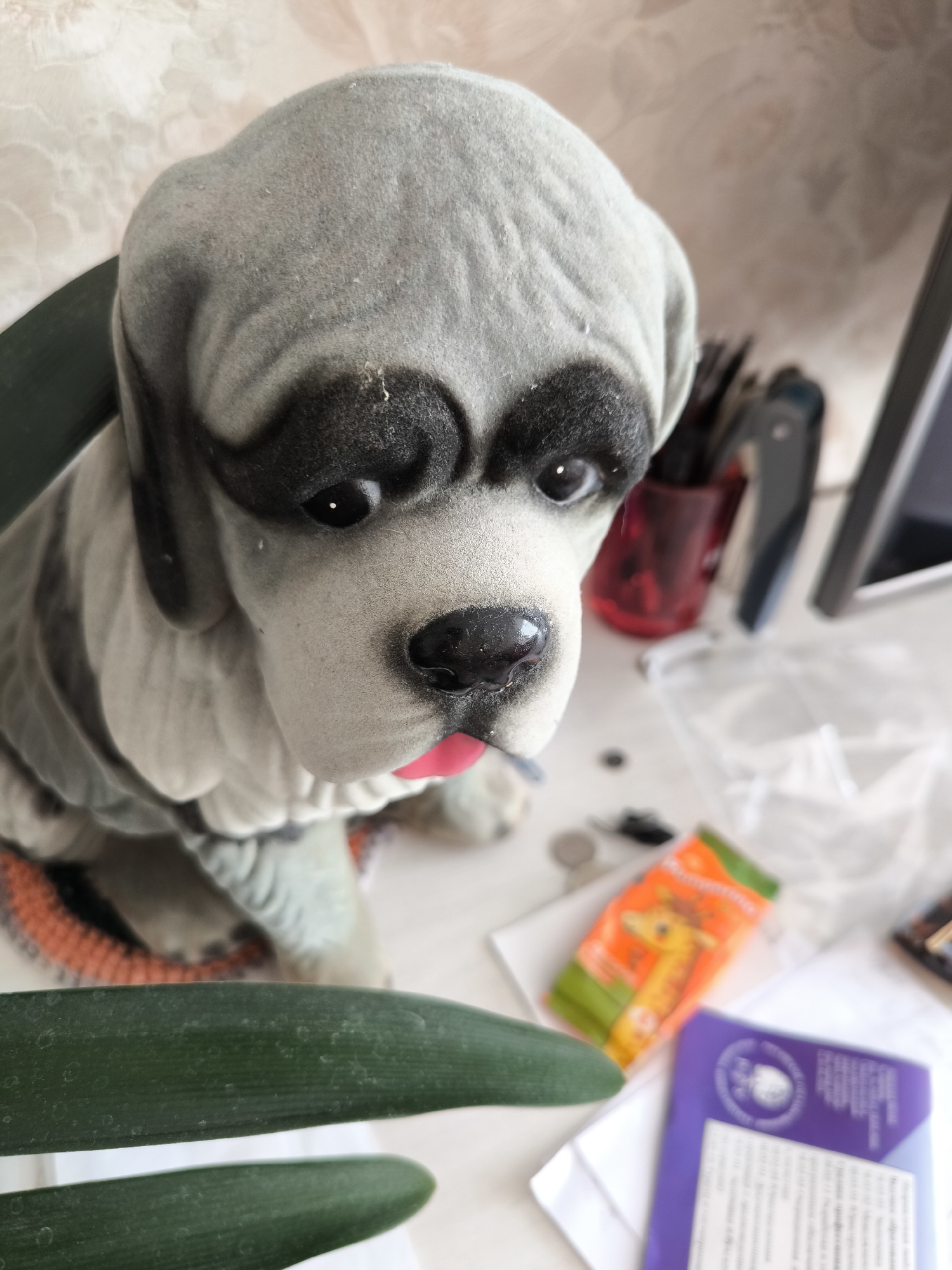 A close-up, high-angle shot shows a stuffed dog toy with gray and black markings, a pink tongue sticking out, and large, sad eyes. The dog is positioned on a light-colored surface, possibly a desk or table, with two large green leaves in the foreground. In the background, various objects are scattered, including a red cup with brushes or pens, a crumpled clear plastic bag, a small orange packet with a picture of a giraffe, a purple card or pamphlet, and a portion of a computer monitor. The background wall has a textured, wallpaper-like pattern. The scene appears to be indoors, likely in a domestic setting in Gal'chino, Russia. There are no people or animals present in the image.