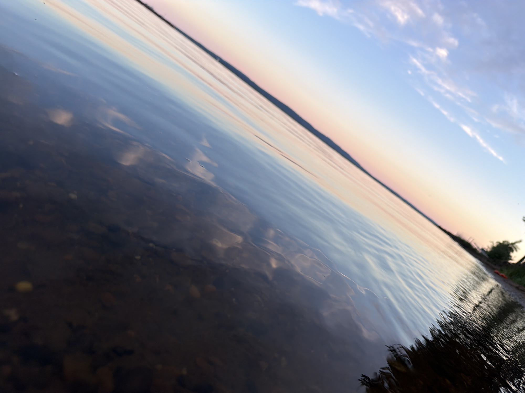 This serene landscape image, taken in Chaikovsky, Russia, captures a vast body of water, likely a lake or large river, under a pastel-colored sky. The photograph is composed at a Dutch angle, emphasizing the expanse of water and sky.
The time of day appears to be either dawn or dusk, indicated by the soft, diffused lighting and the gradual color transition in the sky. Overhead, the sky is a pale blue with scattered, wispy clouds, which gently fades into warm hues of pink, peach, and light orange along the distant horizon.
The foreground is dominated by the water, which is clear enough to reveal the rocky, pebbled bed beneath its surface. The water acts as a mirror, reflecting the soft colors and patterns of the sky and clouds. Gentle ripples disturb the surface, creating a subtle texture and wavy distortions in the reflections. To the right, the immediate shoreline is visible, showing hints of dark earth, sparse vegetation, and a small orange object, possibly a buoy or debris. A dark, indistinct landmass forms the distant horizon line, separating the reflective water from the colorful sky.
The scene conveys a sense of peace and tranquility. There are no people or explicit activities depicted. No text is visible in the image.