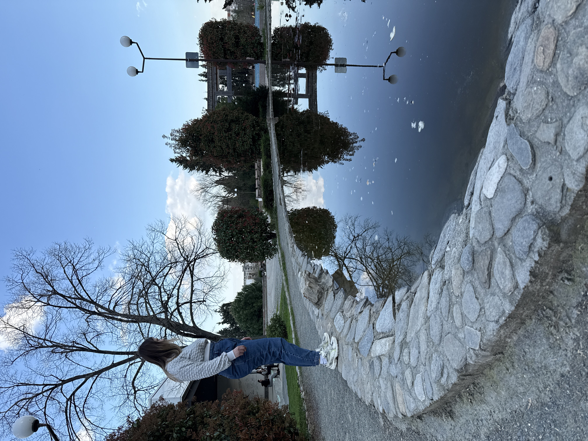 A young woman stands in an outdoor park setting in Stara Zagora, Bulgaria. The woman is positioned along a pathway lined with a stone retaining wall and looks at a water surface which reflects the landscape across. The scene is bathed in daylight, and a blue sky with wisps of clouds is visible. A partially bare tree stands near the young woman, and manicured shrubbery is also present. Street lamps and buildings are reflected in the water.