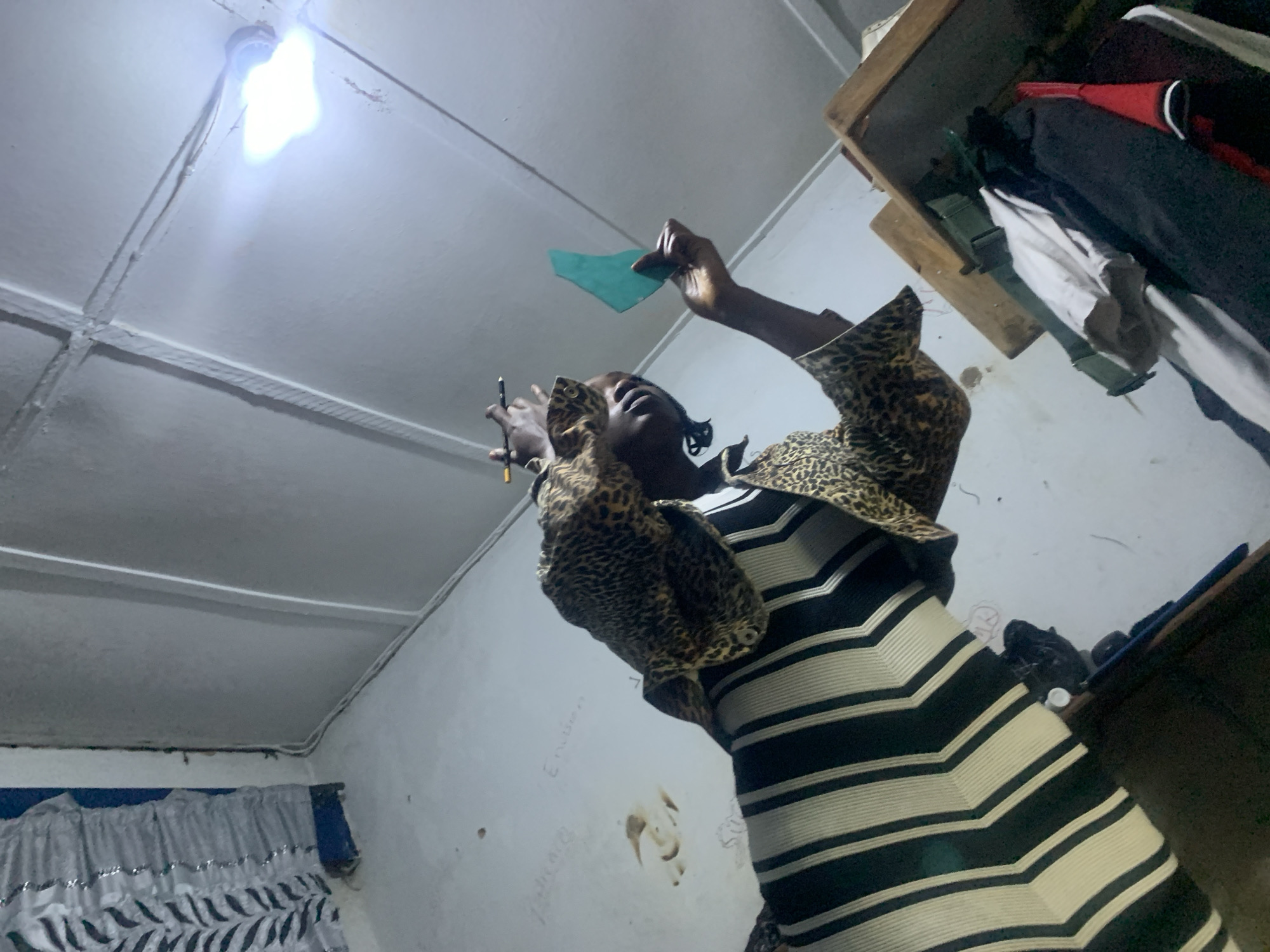 The image shows a person wearing a black and white striped garment and a leopard-print jacket, holding a green object and a pen. The scene appears to be an indoor location in New Stadium, Nigeria, possibly a workshop or living space, characterized by a white-painted ceiling with visible beams and a light bulb. The walls have some markings and what appear to be water stains. To the right, there is a wooden structure with some clothing items stacked on it. The overall lighting is dim, with the overhead light bulb providing the primary illumination.