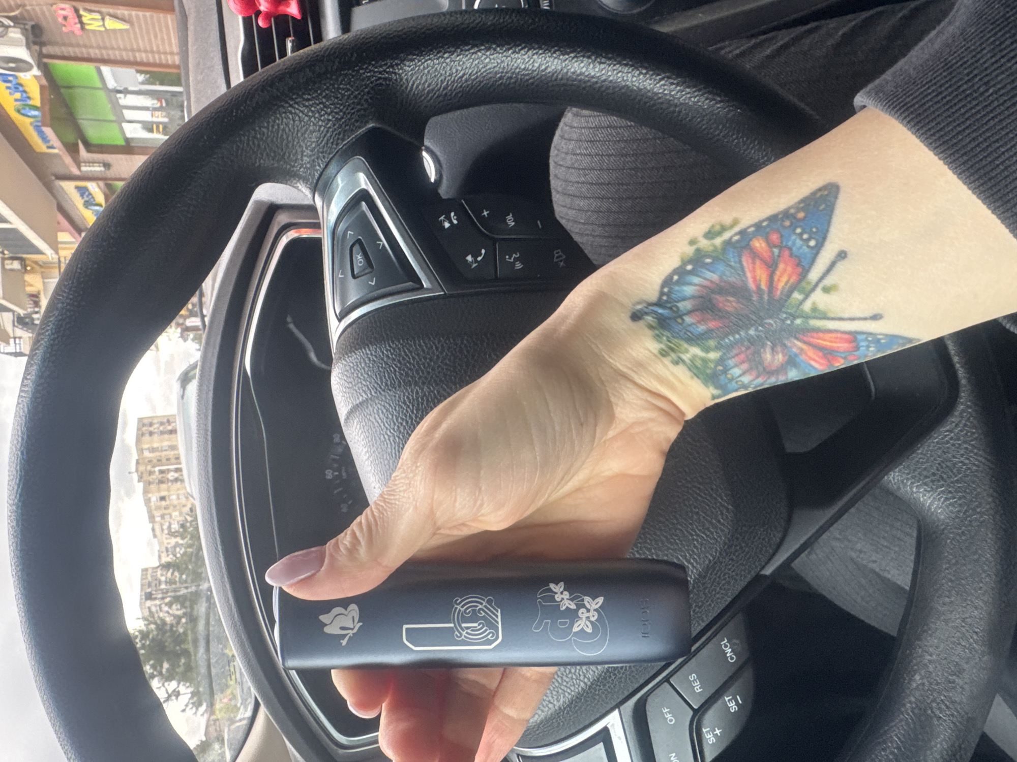 An overhead shot, rotated 90 degrees clockwise, captures a person's arm and hand holding a dark electronic device inside a car. The person, with light skin and light pink/purple nail polish, wears a dark long-sleeved garment. A prominent, colorful butterfly tattoo with blue, orange, and red wings, accented by green, adorns their forearm.
The electronic device held by the person is rectangular and dark grey or black, featuring intricate silver engravings including a butterfly, abstract patterns, and the brand name "SooCii" on one end.
The interior of the car is visible, primarily the black steering wheel with various silver-accented control buttons. On the left, buttons include "OK" and directional arrows. On the right, controls for phone calls, volume ("VOL +", "VOL -"), voice commands, and mode selection are present. Below these, cruise control buttons like "CNCL", "RES", "SET +", "SET -", "OFF", and "ON" can be seen. A small red object, possibly an air freshener, hangs from the dashboard.
Through the car's windshield, a daytime urban street scene is visible. Buildings, some multi-storied, line the street under an overcast or hazy sky. Trees are sparsely distributed. Storefronts with awnings and signs are on the left; visible text on signs includes "NY" or "MV" and "FORD". The overall setting suggests a city environment, consistent with Tbilisi, Georgia as indicated. The person appears to be stationary, holding the device while in the car.