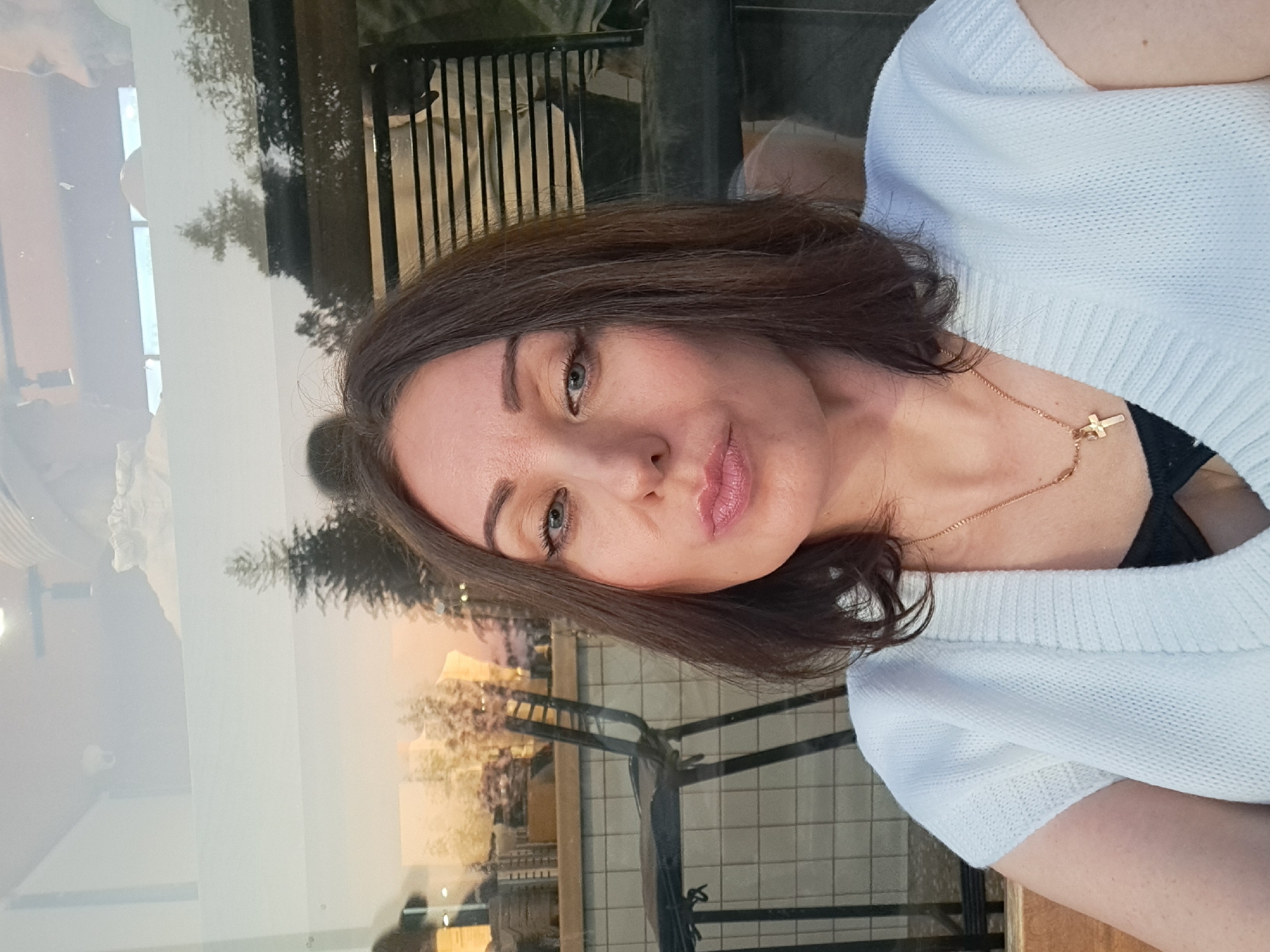 A medium shot captures a woman with shoulder-length dark brown hair and fair skin. She is looking directly at the camera, her head slightly tilted. She has blue eyes, defined eyebrows, and is wearing subtle makeup. Her lips are slightly pursed, and she is wearing a gold necklace with a cross pendant over a black bra and a white knit cardigan. The background appears to be a cafe with large windows reflecting the outside, showing trees and outdoor seating. The overall lighting suggests it is daytime, possibly late afternoon. The scene is composed in a way that suggests a casual selfie taken within an urban setting, likely a public establishment. The specific location of Irkutsk, Russia, is not discernible from the visual information provided in the image.