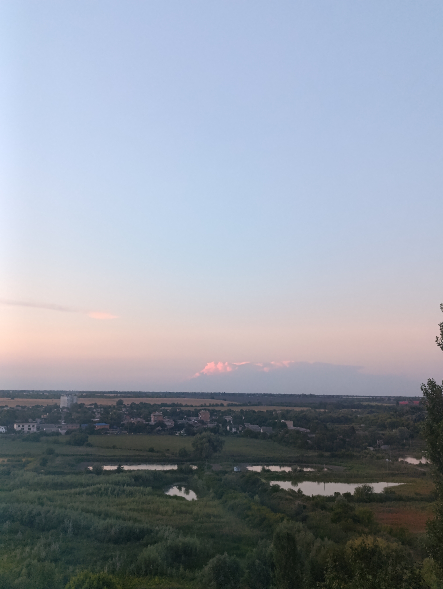 An aerial landscape photograph captures the city of Bila Tserkva, Ukraine, at dusk. The sky transitions from a pale blue at the top to a soft pink and orange gradient near the horizon, with a few wispy clouds tinged pink. Below the sky, a panorama of the city unfolds, featuring a mix of low-rise buildings and some taller structures, interspersed with lush green trees and fields. Several bodies of water, appearing as small lakes or ponds, are visible within the landscape, reflecting the soft light of the evening sky. The foreground is dominated by dense, dark green foliage, suggesting the vantage point is from a higher elevation, possibly overlooking a park or natural area on the outskirts of the city. The overall scene conveys a peaceful, rural-urban transition during twilight.