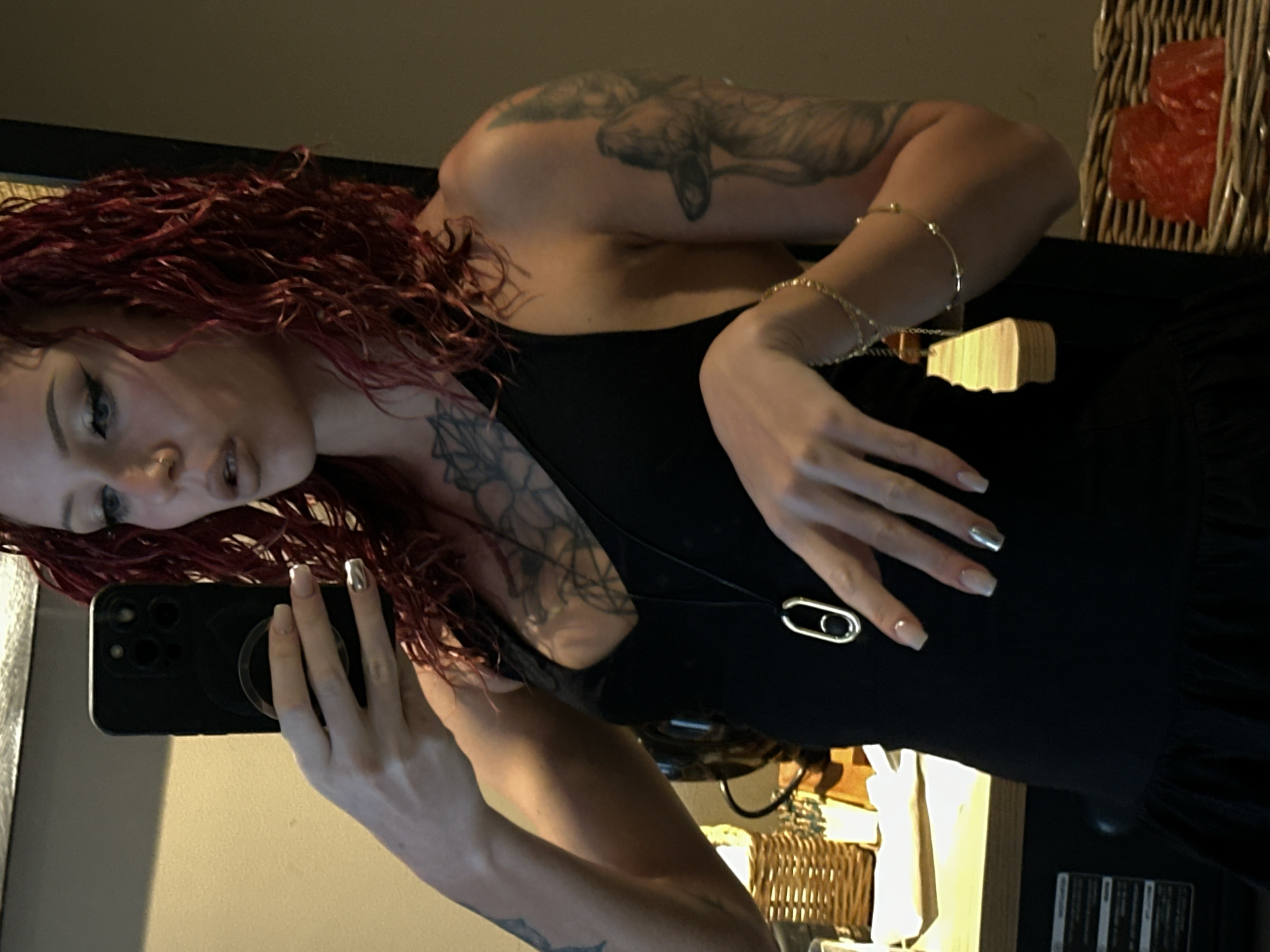 A woman with red curly hair, tattoos on her arms and chest, and manicured nails takes a selfie using a black smartphone. She wears a black top with a pendant necklace and gold bracelets. The background shows a basket with wrapped items, a wicker basket, and other household items. The location is in a room. The city of Unknown, Indonesia is not relevant to this scene.