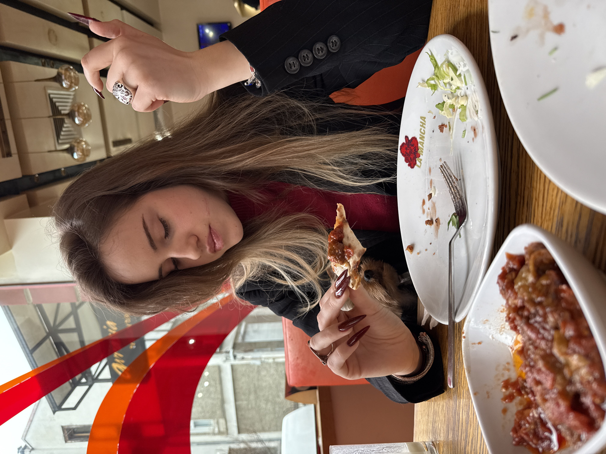 A young woman is seated at a table in a restaurant, possibly in Sofia, Bulgaria, based on the "LA MANCHA" logo on a plate, potentially referring to a local eatery. The woman, with long brown and blonde hair, is dressed in a red turtleneck sweater and a black blazer. She has manicured nails and several rings on her fingers. She holds a piece of food in her right hand, while her left hand is raised, possibly gesturing. A small dog is also present, partially visible near her left hand. The table is covered with used dishes, including plates with food remnants and a small bowl containing a tomato-based dish. A glass of water is also on the table. The background shows a window with a building outside.