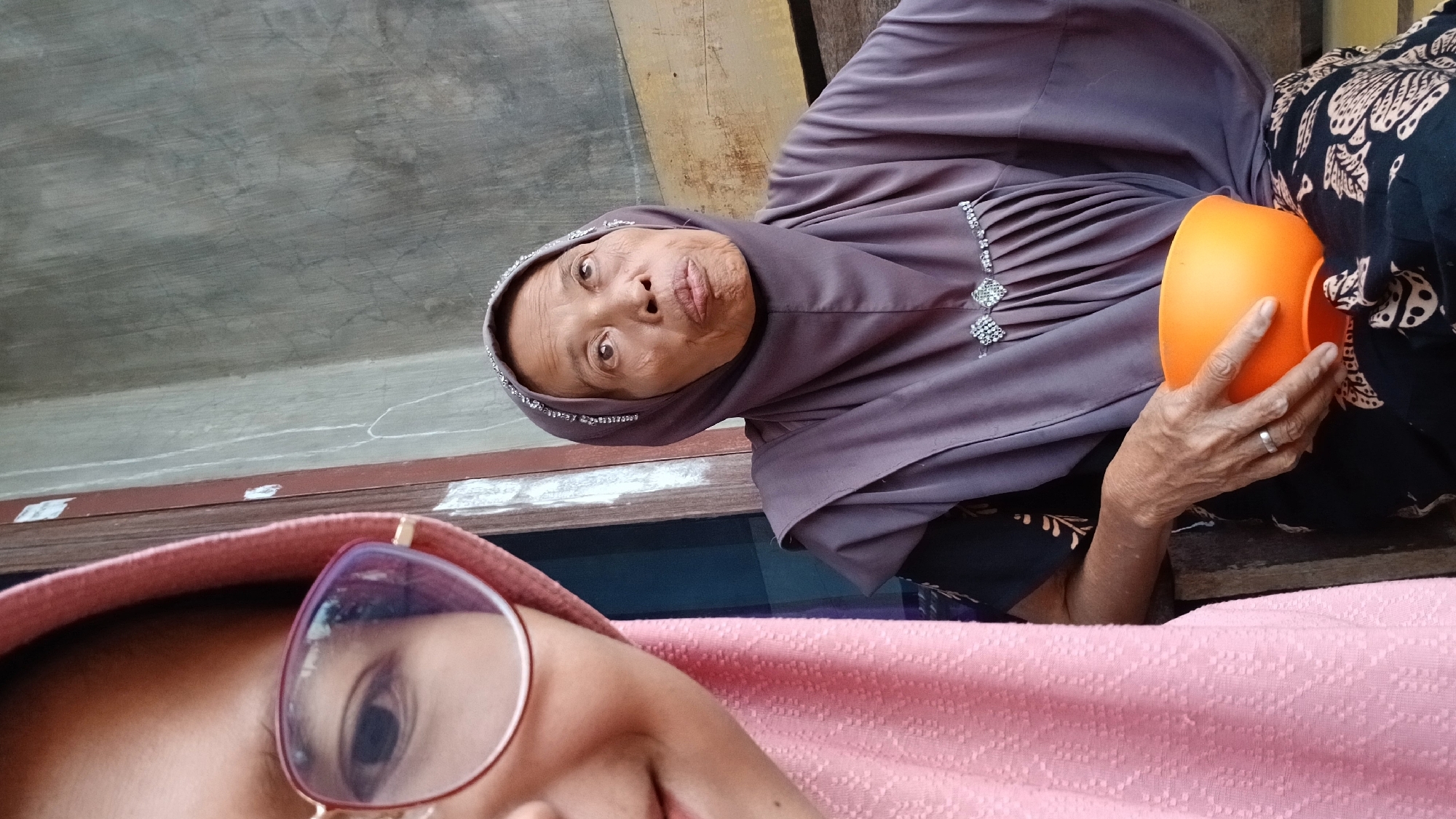 In Lhokseumawe, Indonesia, two individuals are visible. One person, wearing a mauve hijab adorned with decorative embellishments, is holding an orange container. This individual's face is turned upwards toward the camera. The other person, partially visible in the foreground, is wearing eyeglasses and a pink head covering. The environment includes what appears to be a concrete wall and wooden planks, suggesting an outdoor setting.