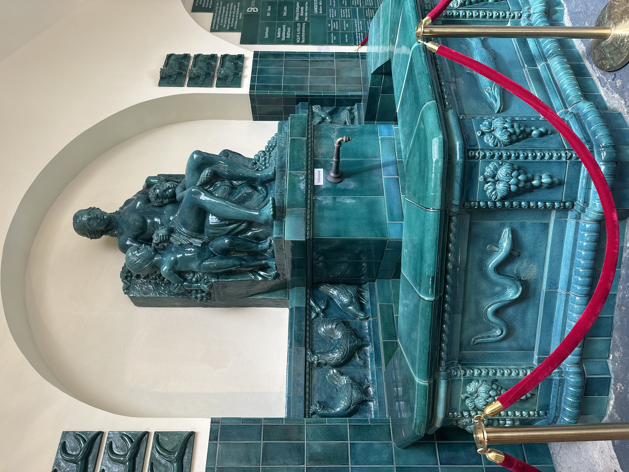 The image shows a turquoise tiled fountain located in Stuttgart, Germany, specifically at the entrance of the Markthalle. The fountain features a statue of a woman with a child on her lap, carved in the same turquoise material. Reliefs of roosters, grapes, a snake, and fish are also incorporated into the fountain's design. A red velvet rope barrier is in place in front of the fountain. Several green tiled plaques with text are mounted on the wall behind the fountain, listing sponsors and information about the Markthalle. The overall environment appears to be an indoor, arched entryway.