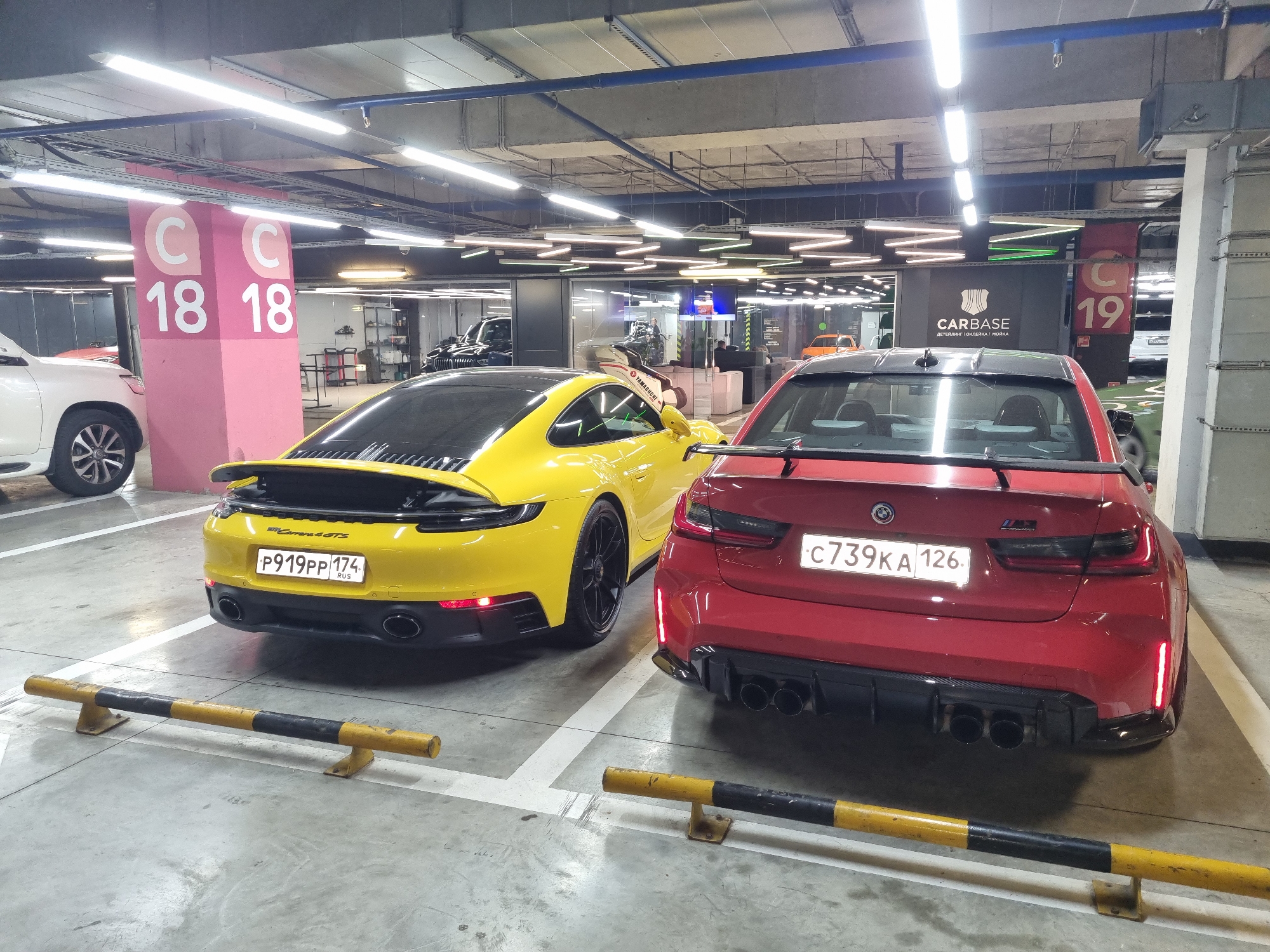 This media file captures a ground-level view within an artificially lit underground parking garage in Sochi, Russia. The central focus is on two high-performance sports cars parked side-by-side: a bright yellow Porsche 911 Carrera GTS and a red BMW M3.
The yellow Porsche, seen from its rear-left, prominently displays "Carrera GTS" on its back and a Russian license plate "P919PP 174 RUS". Its sleek design includes twin exhaust pipes and black multi-spoke wheels.
To its right, the red BMW M3 is shown from the rear, featuring a distinctive quad exhaust system, a small rear spoiler, and an aggressive diffuser. Its Russian license plate reads "C739KA 126". Both cars are parked behind yellow and black striped car stoppers, which delineate the parking spots.
In the background, a white SUV, possibly a Toyota Land Cruiser, is partially visible to the left. To the right, through glass walls, a car service or detailing center named "CARBASE" is discernible. The "CARBASE" sign, written in Cyrillic, also states "ДЕТЕЙЛИНГ | ОКЛЕЙКА / МОЙКА", indicating detailing, wrapping, and washing services. Inside this facility, other vehicles, including a dark SUV and an orange car, can be seen.
Concrete pillars marked "C 18 18" and "C 19" indicate parking section numbers. The ceiling features exposed pipes, conduits, and numerous fluorescent strip lights, typical of a modern underground parking facility. No people are visible, and the scene appears to be static, with the cars simply parked.