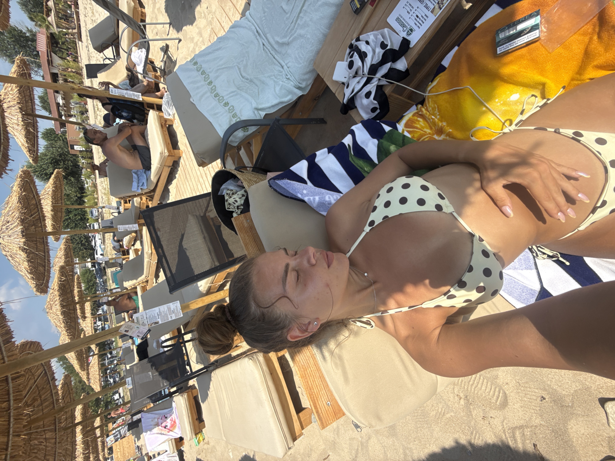 At Ofrynio Beach, Greece, a woman in a polka-dot bikini lies on a sun lounger. She has her eyes closed and her hand resting on her stomach. The beach is lined with sun umbrellas and loungers. Several other people are visible in the background, some relaxing on loungers and others walking. The sand is light-colored, and the water is not visible in the frame. The scene appears to be during the day, with bright sunlight casting shadows.