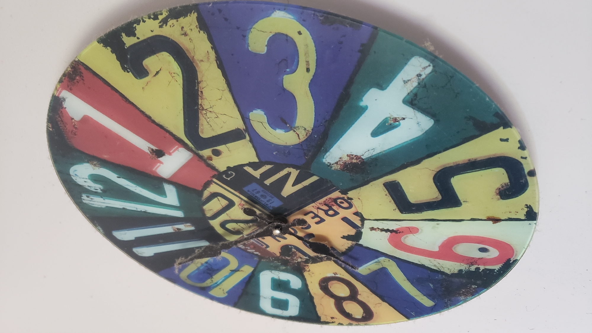 The image displays a clock face made from what appears to be a collage of license plates. The clock is circular and features large numbers from 1 to 12, each set against a different colored background derived from the license plates. The hands of the clock are black and point to approximately 10:58. The clock is mounted on a white wall. There is no indication of the city of Bratislava, Slovakia in the image.