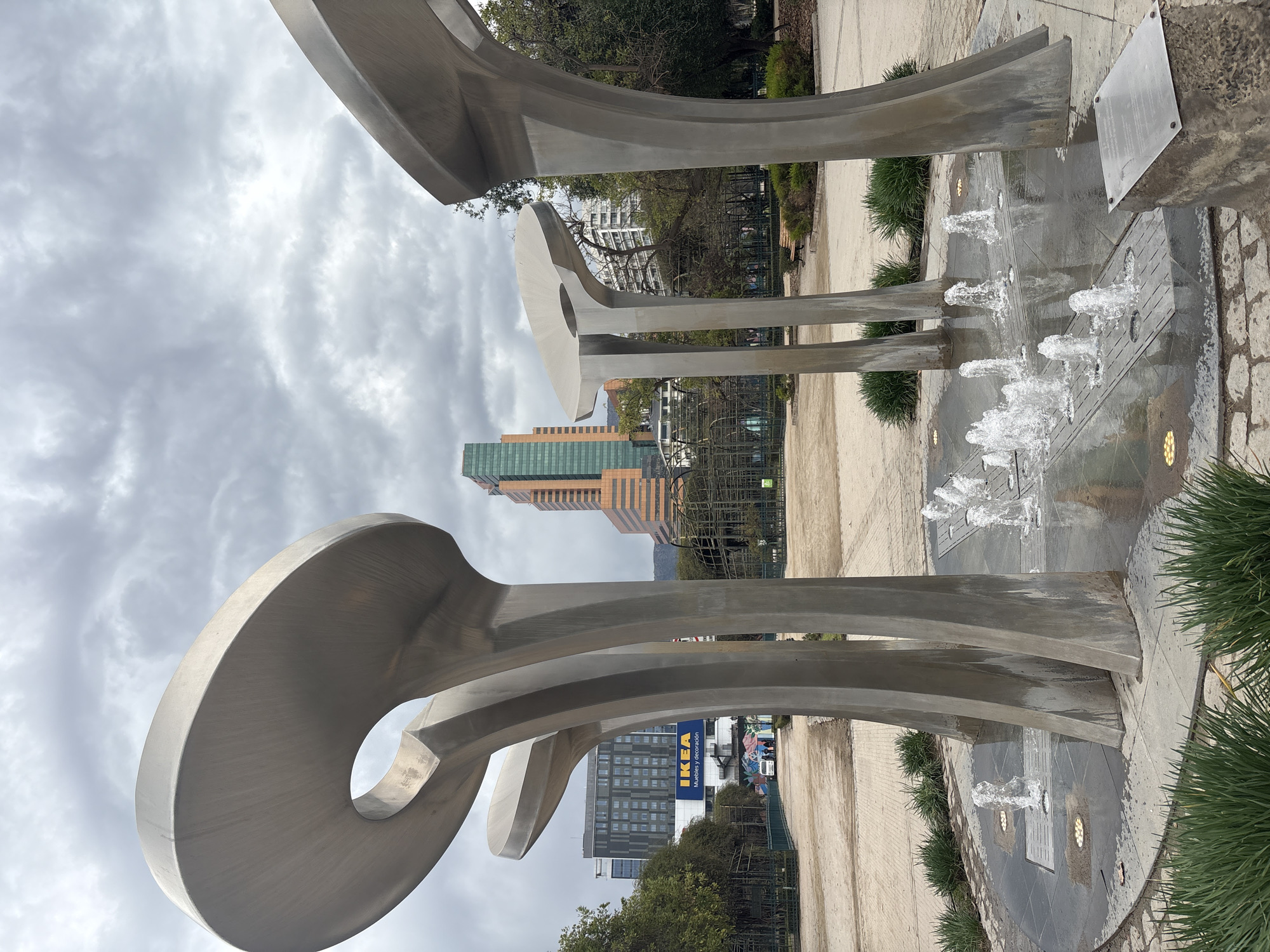 An outdoor view captures a contemporary metal sculpture with multiple curved, mushroom-shaped elements supported by thick, columnar legs, situated within a fountain. Water jets spray upwards from the fountain's surface, creating a dynamic visual against the backdrop of a city park. In the background, modern high-rise buildings stand tall, including a prominent IKEA store with its distinctive blue and yellow signage. The sky is overcast with dramatic clouds, suggesting a changeable weather day. The park features paved pathways, scattered benches, and a metallic trellised structure, indicating a public urban green space. The sculpture and fountain are the central focus, with the urban environment providing context. This scene is located in Parque de las Esculturas in the Providencia commune of Santiago, Chile.
