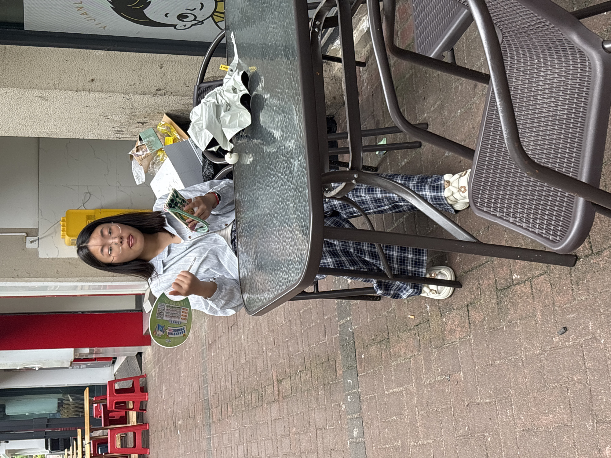 A person is seated at an outdoor table in Jiangjin, China. The person is wearing glasses, a light-colored, button-down shirt, and plaid pants. They are holding a hand fan in one hand and what appears to be a mobile phone in the other. The table is made of glass with a metal frame. There are metal chairs around the table. In the background, there is a building with a red door and stacked red stools. There's also an obscured shop sign at the top of the frame. The ground is paved with bricks. A discarded umbrella and other items sit on the table next to the person.