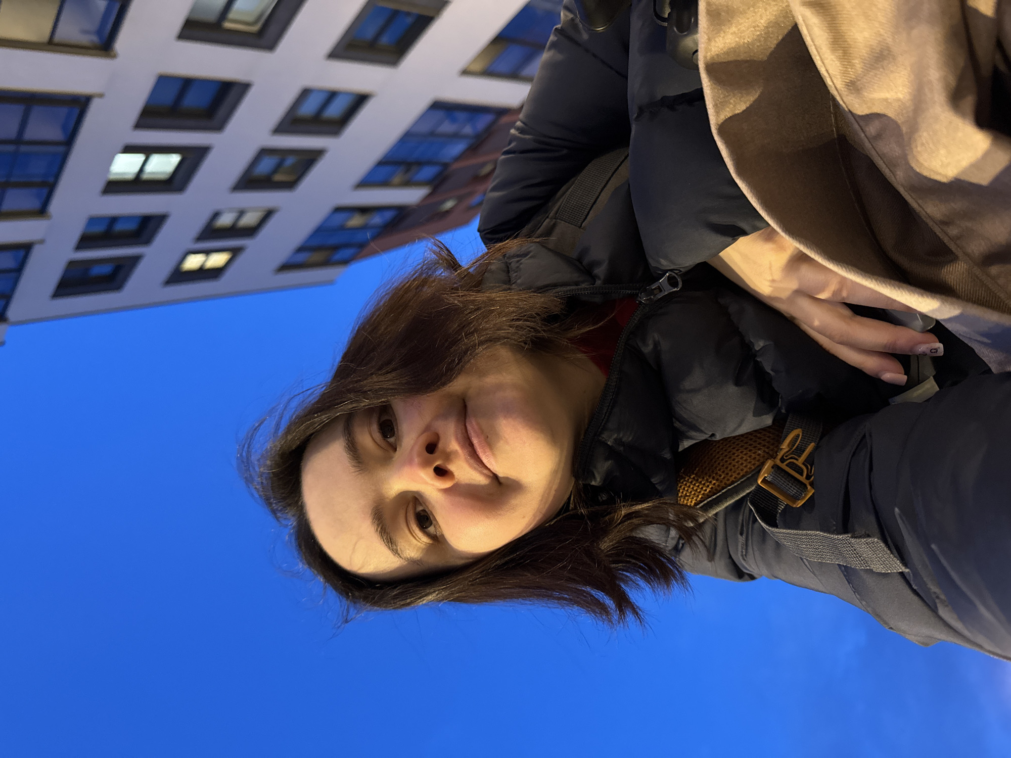 A person takes a selfie with a building and blue sky in the background. The building has multiple windows, some of which are illuminated. The person is wearing a dark puffer jacket and a light-colored bag. The setting appears to be outdoors, likely an urban area. The specific location in Izhevsk, Russia, is not discernible from the image.