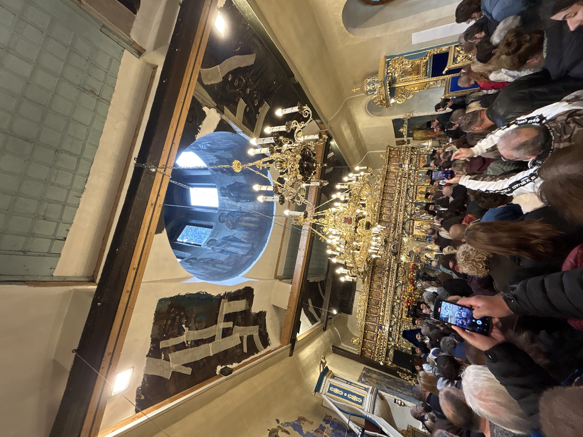 The image depicts the interior of an Eastern Orthodox church in Asenovgrad, Bulgaria, observed from within a dense congregation looking towards the altar. A prominent, multi-tiered, ornate golden iconostasis dominates the central background, featuring numerous icons and religious figures. Behind the iconostasis, several clergy members in black vestments are visible, with one figure in elaborate golden vestments appearing further back near the altar.
Above the iconostasis, a large, ornate golden chandelier with multiple lit candles or bulbs is suspended from the ceiling. Further up, a circular opening reveals a dome or cupola with windows letting in natural light and frescoes depicting religious figures. The surrounding ceiling and upper walls exhibit painted frescoes, some areas of which show signs of age, flaking paint, or restoration efforts indicated by white patches and exposed wooden beams. A section of the ceiling to the top right features a light blue, coffered pattern. Additional, smaller square recessed lights are visible in the ceiling.
To the left of the iconostasis, a blue and gold ornate pulpit or stand is visible. To the right, another ornate blue and gold structure, possibly a bishop's throne or another pulpit, stands against a light-colored arched wall.
The foreground is filled with a standing crowd of individuals, varied in age, predominantly facing the iconostasis. Numerous members of the congregation are holding up smartphones, actively recording or photographing the religious service. One individual in the immediate foreground prominently displays a black smartphone capturing the scene. The overall scene conveys a religious gathering in a historically rich church interior.