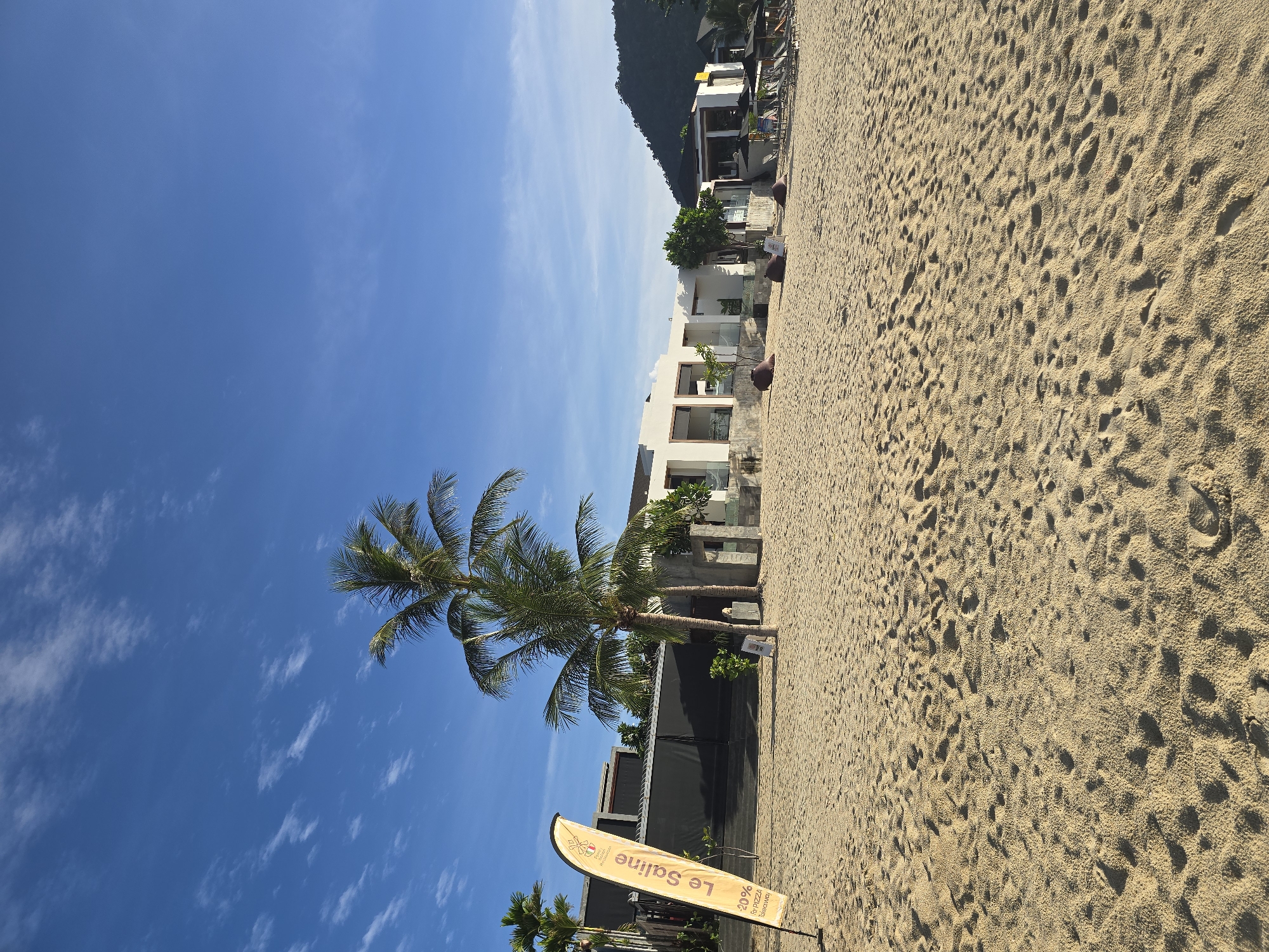 The image depicts a beachfront with a modern resort building in the background. The foreground is dominated by a sandy beach with footprints. In the mid-ground, a tall palm tree stands to the left of the resort, and a yellow flag with text is planted in the sand to its left. The resort features a white, multi-story structure with large windows and balconies, partially obscured by vegetation. Several lounge chairs and umbrellas are visible on the beach in front of the resort. The sky is a clear blue with scattered white clouds. A hillside covered in greenery is visible in the far background.