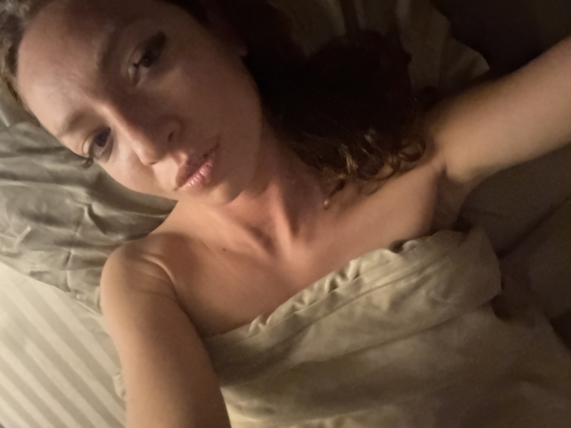 A woman lies in bed, covered by a blanket. She is facing the camera, with her head tilted. She has brown hair and appears to be wearing makeup. The sheets are striped. The location in the city of Hà Giang 1 Ward, Vietnam, is not determinable from the image.