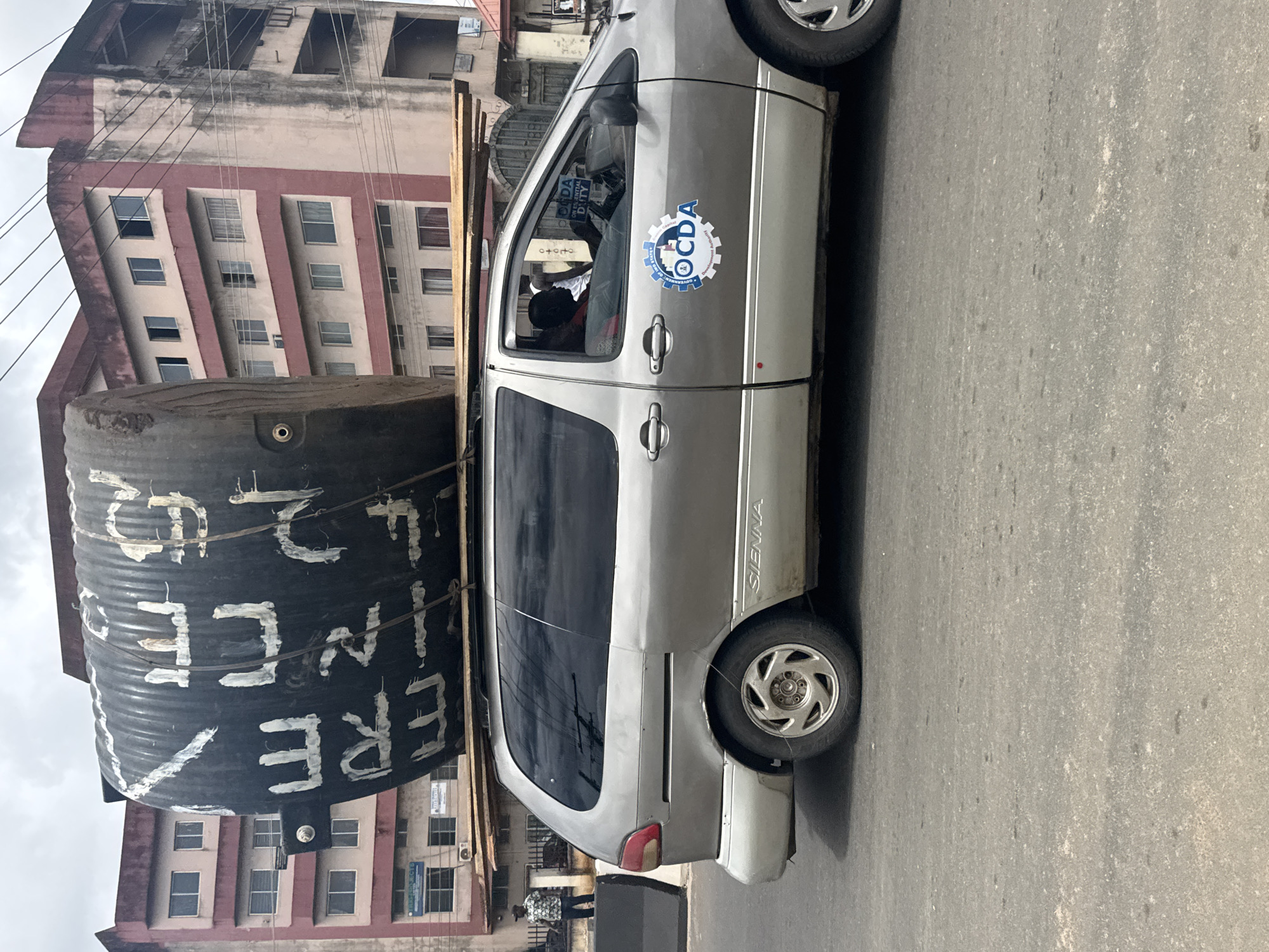 A side view of a silver minivan is captured on a street in Owerri, Nigeria, with a large, black cylindrical object secured to its roof with wooden planks. The cylindrical object appears to be a water tank with white lettering. A man is visible driving the minivan. The street is paved, and buildings line the background. A person in a patterned shirt stands on the sidewalk to the left of the vehicle. The sky is overcast.