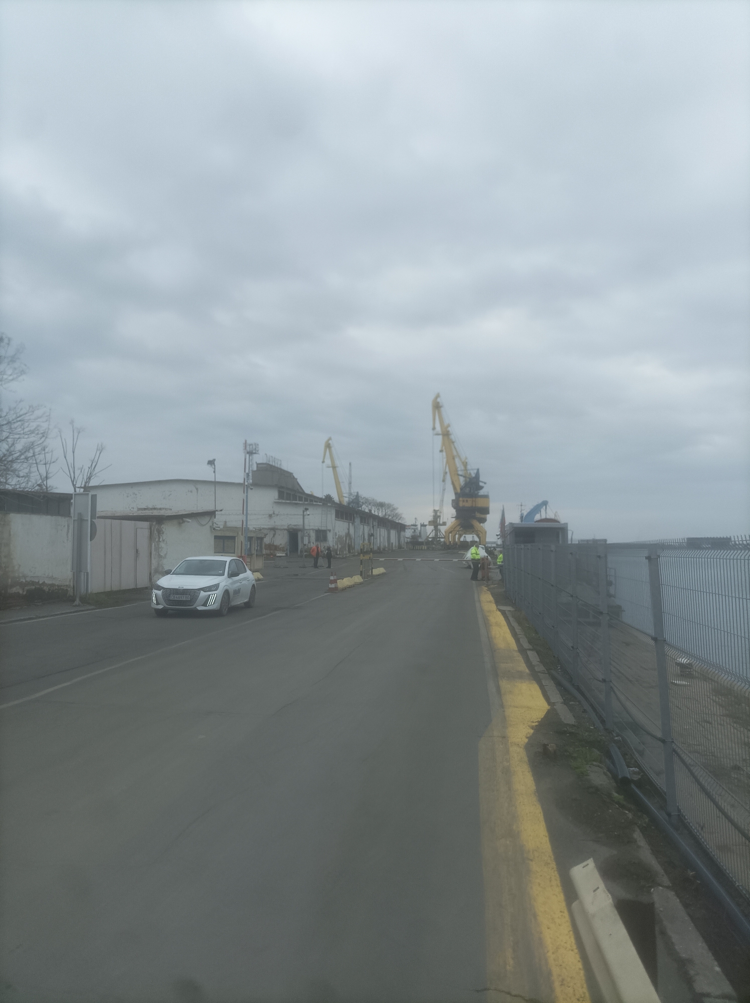 The image depicts a street scene in the port of Burgas, Bulgaria. The sky is overcast. A white car drives along the street. Several yellow port cranes are visible in the background, as well as industrial buildings. Along the right side of the street, there is a fence, and two people in yellow vests are visible.