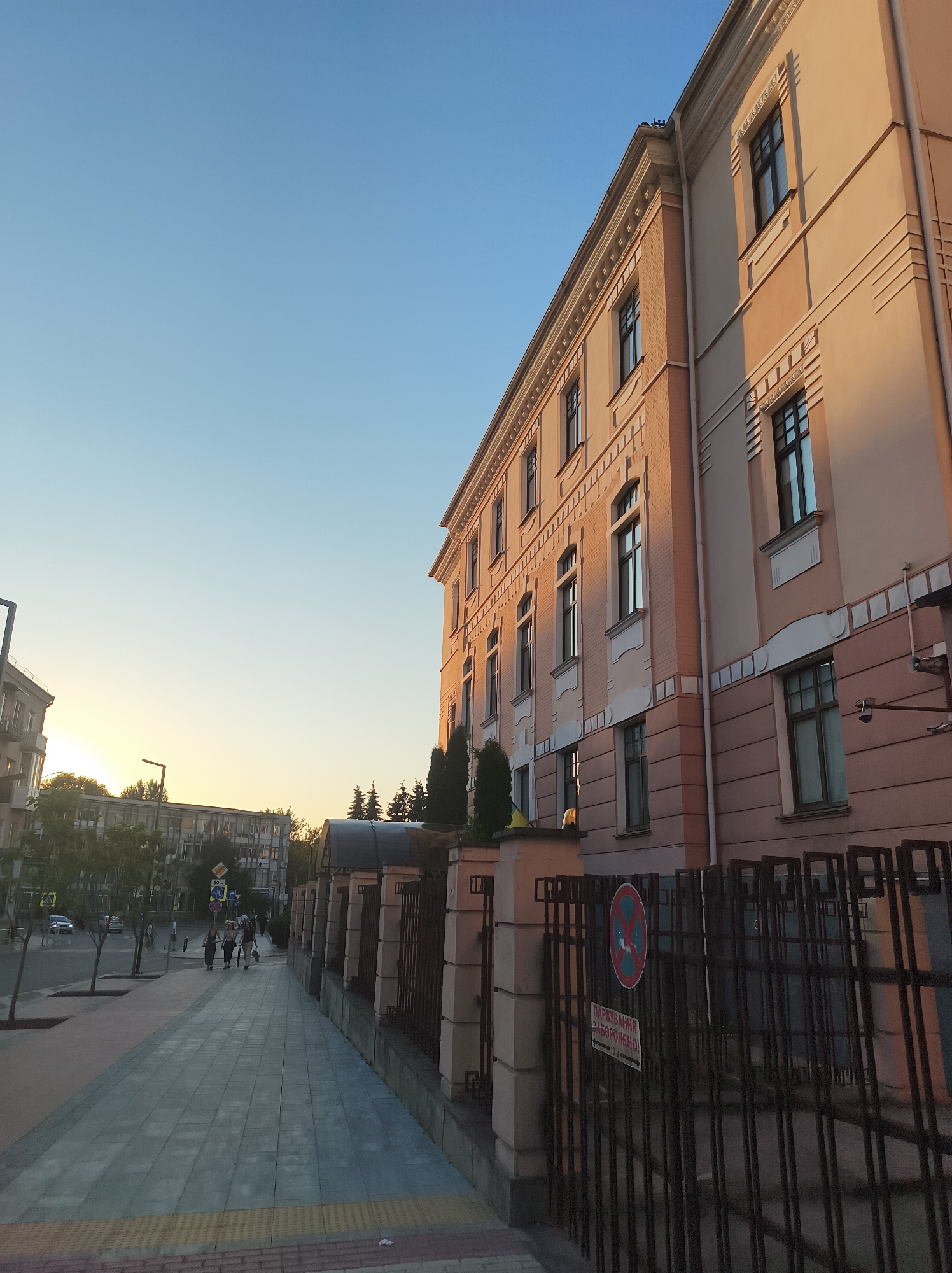 The image depicts a street scene in Ternopil, Ukraine, during what appears to be late afternoon or early evening, given the warm light. On the right side of the frame, a large, light-pink building with multiple windows and decorative architectural details dominates the scene. The building has a classical or early 20th-century style. In front of the building, a dark metal fence runs along the sidewalk, with a circular red and blue sign prohibiting parking and a rectangular sign with Ukrainian text.
To the left of the building, a wide pedestrian sidewalk paved with grey tiles extends into the distance. Several trees are planted along the edge of the sidewalk, casting shadows. In the background, across the street, other buildings are visible, including a multi-story building with numerous windows and air conditioning units, suggesting it might be an office or residential complex.
Several people are walking on the sidewalk in the middle ground, appearing as small figures. A few cars are visible on the street in the background. The sky above is clear and a soft blue, transitioning to a lighter yellow hue near the horizon on the left, indicating the setting sun. The overall atmosphere is calm and urban.