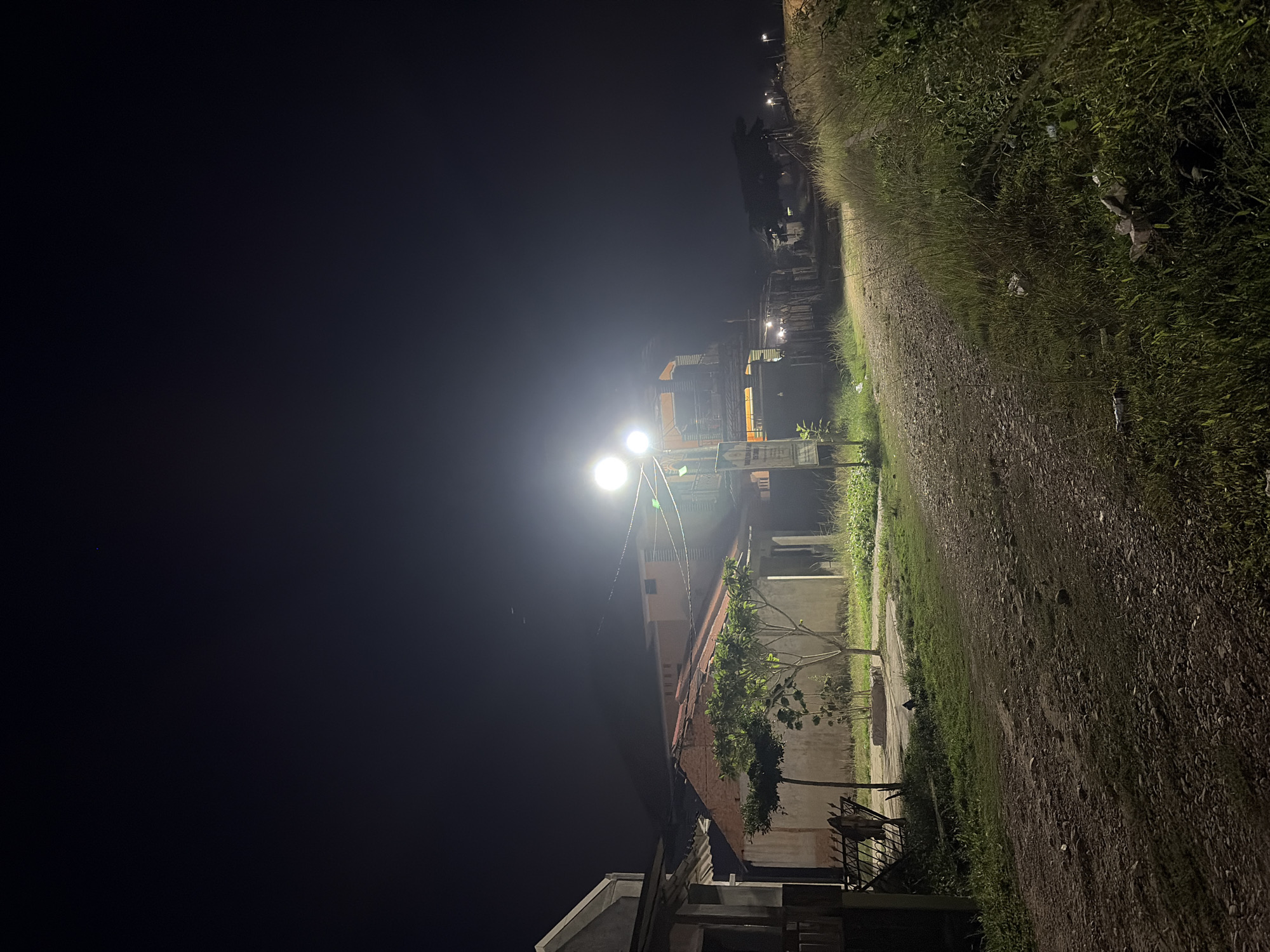 The image captures a nighttime scene on a dirt road in Tupah, Indonesia. Two bright streetlights illuminate the area, casting a glow on a small tree with green foliage situated in the foreground, to the left of the road. Adjacent to the tree, a house with a red-tiled roof is visible. Further down the road, other buildings and structures can be seen, also lit by streetlights. The road itself is composed of dirt and small stones, with grass and vegetation growing along its edges. The sky above is dark and clear, with a single small blue light point visible in the upper portion of the image. There are no people present in the scene.