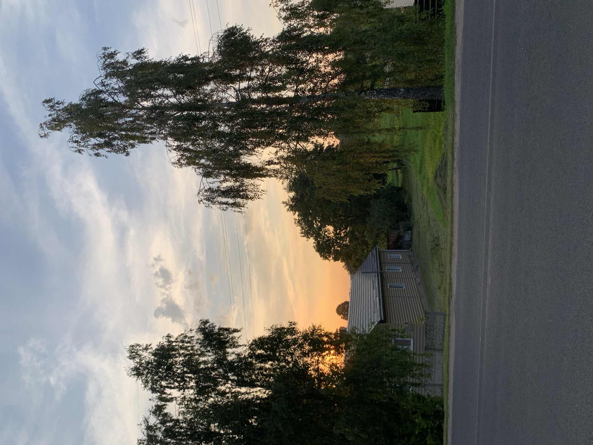The image depicts a rural scene in the city of Kapyĺ, Belarus, during sunset. A paved road runs horizontally across the foreground. To the left of the road, a long, low wooden house with a dark roof is visible, partially obscured by trees. The house has several windows with white frames. A low, decorative concrete fence runs in front of the house.
Behind the house and fence, a grassy area slopes upwards, dotted with trees and foliage. A large birch tree with weeping branches stands prominently in the middle ground, casting a shadow. The sky above is a gradient of soft blues and oranges, with scattered clouds, indicating the setting sun. The sun itself is visible peeking through the leaves of trees on the far left, casting a warm glow. Electrical wires are strung across the upper portion of the image. The overall impression is one of a tranquil, rural evening. There are no people or animals visible in the image.