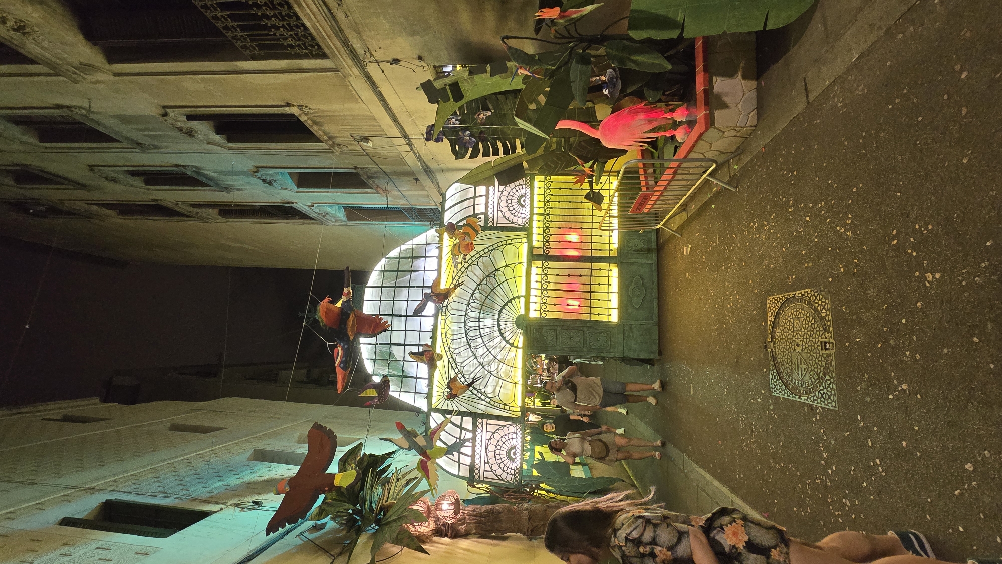 The image captures a narrow alleyway at night, possibly in the El Born neighborhood of Barcelona, Spain, judging by the architectural style and the presence of what appears to be a Barcelona event barrier. The alley is illuminated by artificial lights, casting a warm yellow and green glow. The walls of the buildings flanking the alley are made of stone and have multiple windows. Suspended above the alley are colorful, decorative bird figures.
In the foreground, a woman with long dark hair is partially visible on the left, wearing a floral print dress. Further down the alley, two couples are walking towards the camera. The group on the left consists of a man and a woman, both casually dressed. The couple on the right are also casually dressed.
The alley opens up to a large arched gateway with ornate metalwork and illuminated with bright yellow lights, suggesting an entrance to a venue or event. In front of the gateway, there's a section with large green foliage decorations and a bright pink flamingo statue, adding a tropical theme to the scene. A metal barrier, marked with "Barcelona," is set up to the right of the flamingo. The ground is paved and appears to be slightly wet, with scattered light-colored debris. A patterned manhole cover is visible in the center of the alley.