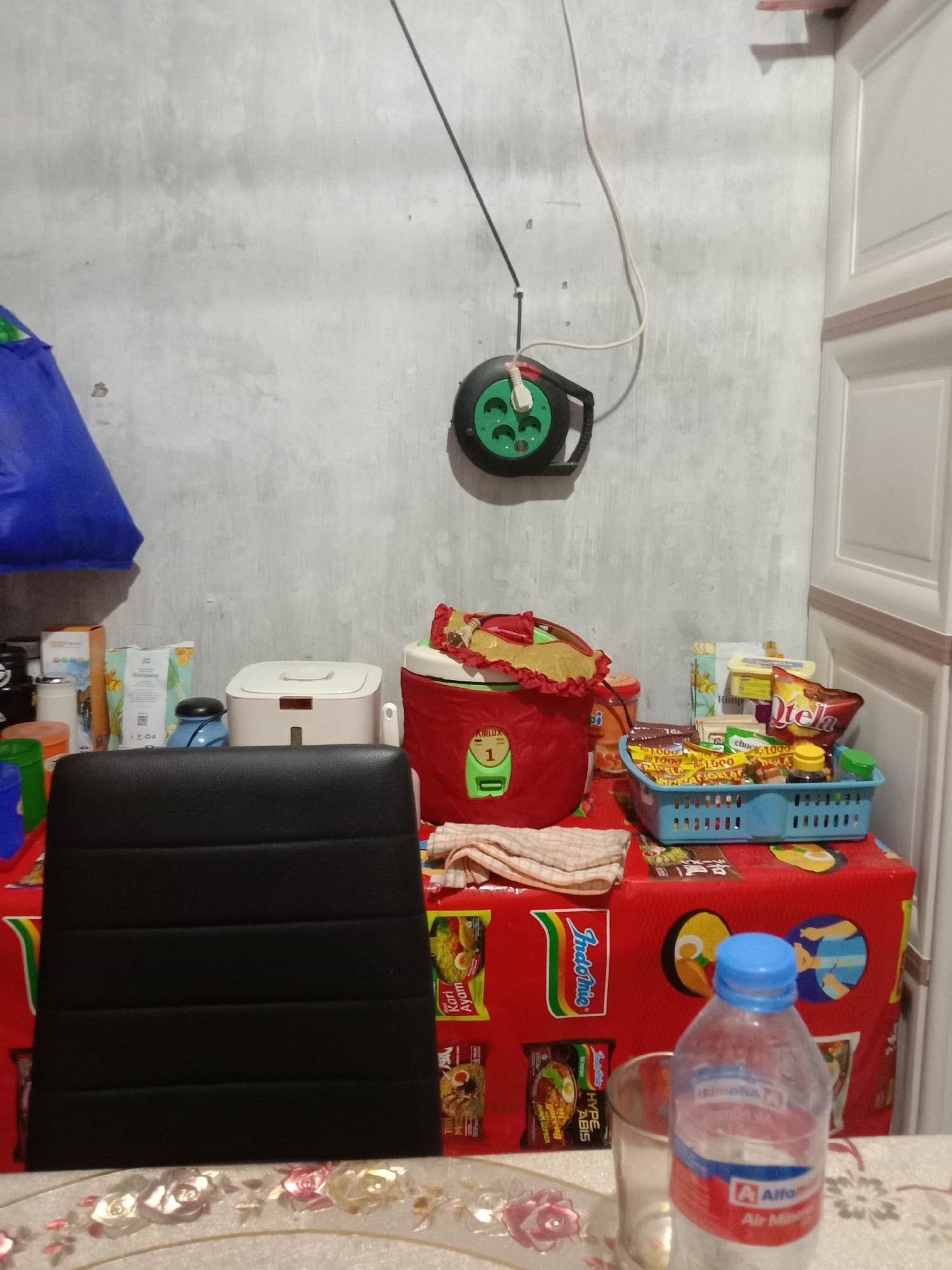 The image depicts an indoor scene, likely a kitchen or dining area, with various household items organized on a red-covered table. The table is adorned with food items, including packaged snacks and instant noodles, arranged in a blue basket and scattered around. A red rice cooker is prominently placed on the table, accompanied by a white towel draped over it. To the left, a black office chair is positioned in front of the table. Behind the table, a white electrical extension cord reel is mounted on a textured gray wall, with a black cord extending from it. A blue plastic bag is visible on the left side of the frame. The overall environment suggests a functional living space. This scene is located in the city of Kwandang, Indonesia.